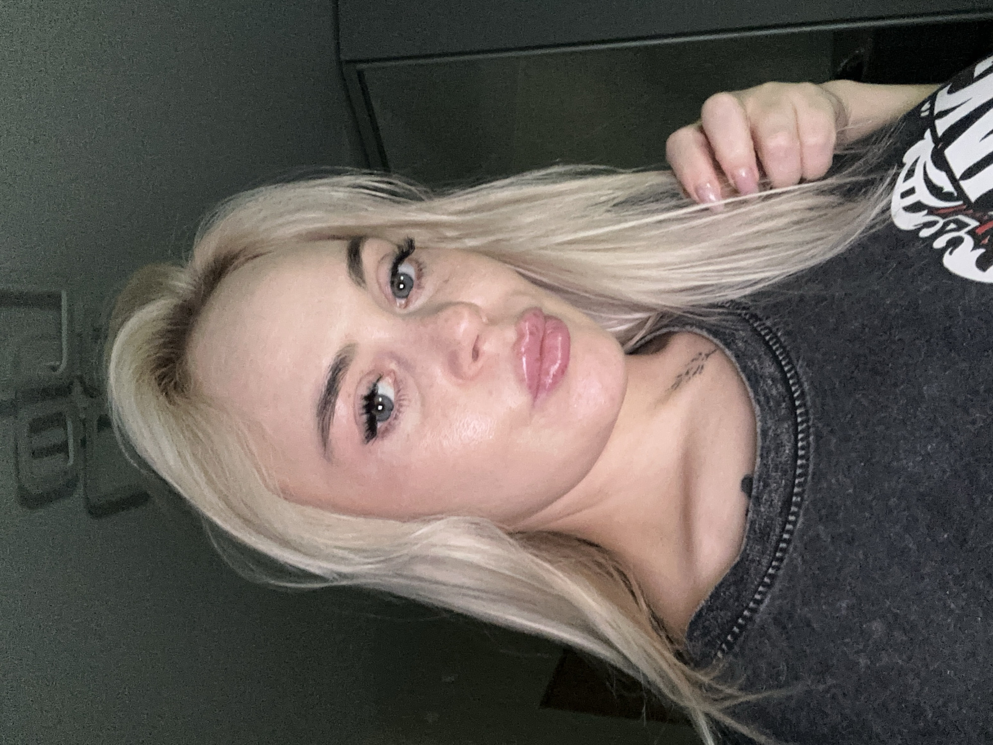 The image is a close-up, tilted selfie of a woman with long, straight blonde hair and light-colored eyes (appearing blue or grey). Her face is central, with defined eyebrows, dark eyelashes, and full, glossy lips pursed in a "duck face" or kissy expression. She has clear skin with a subtle sheen.
Her right hand, adorned with light pink, manicured nails, is visible on the upper right, gently holding a section of her blonde hair. She is wearing a dark grey top, which appears to be a sweater or t-shirt, featuring a visible textured seam or zipper detail along the neckline/shoulder. Part of a white and black graphic design, possibly an abstract or skeletal pattern, is visible on the upper right side of her shirt. A small, dark tattoo, with illegible text, is visible on her left collarbone/neck area, accompanied by a small dark mole or mark.
The background is dark, out of focus, and indistinct, suggesting an indoor setting with subtle structural elements that are difficult to identify. The lighting appears to be somewhat dim, highlighting her face. The overall activity is a casual, posed selfie. No legible text is visible in the image.