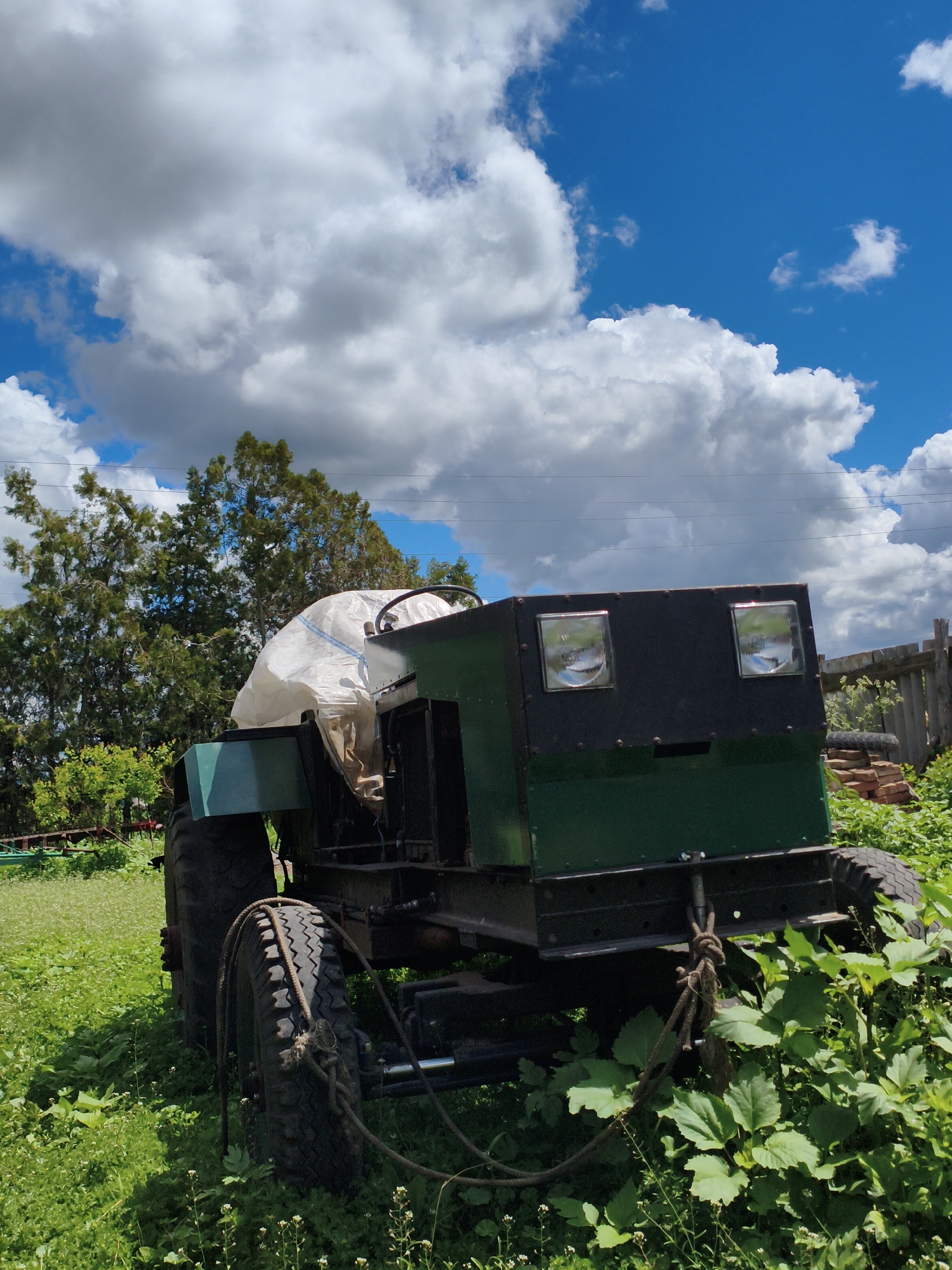 The image depicts a green and black homemade tractor in an overgrown field, likely in a rural area. A white, full sack covers the driver's seat. The tractor is partially obscured by green vegetation. A rope is tied to the back of the tractor and around the rear tire. In the background, there are trees and a blue sky with white clouds. Based on the prompt, the location is Ustyvytsia, Ukraine.