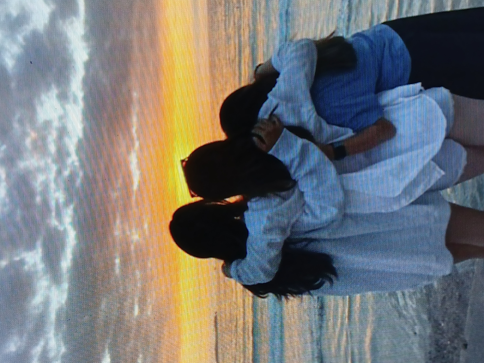 Three people stand in a group, embracing in front of a sunset over the ocean. The person on the right wears a blue top and dark bottoms. The two people on the left are wearing light-colored tops and bottoms. The location appears to be a beach, with the ocean and sunset in the background. While it may be from Juan Griego, Venezuela, this cannot be determined without further information.