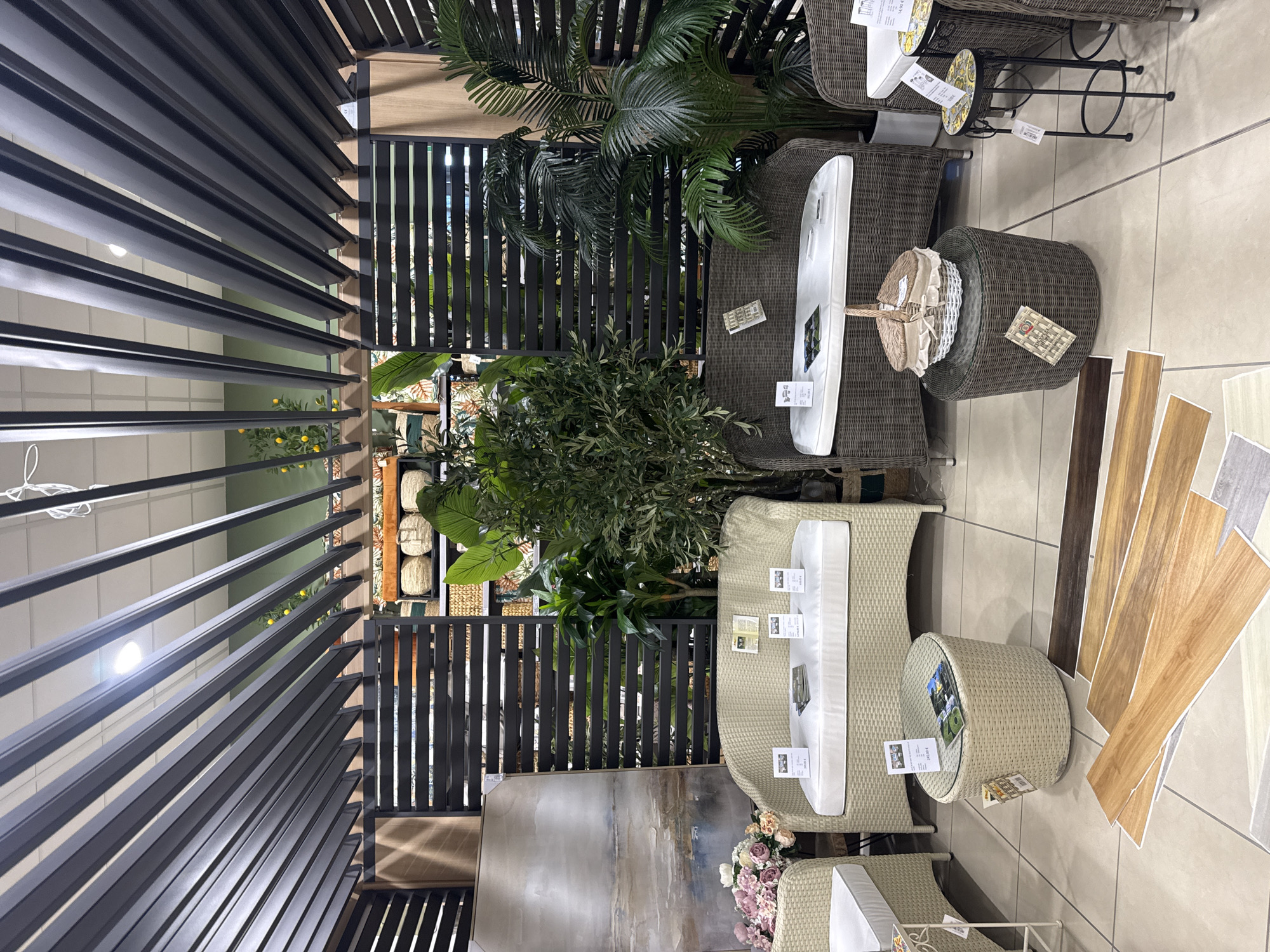 This appears to be a showroom display of outdoor furniture, potentially inside a store. The scene includes at least two sets of wicker-style patio furniture with white cushions. There are also round wicker tables, plants (including a palm), and what appears to be a piece of artwork displayed. Samples of flooring materials are arranged on the floor.
Given the presence of pricing tags and display materials, it is located in a retail or showroom environment, specifically in Pomezia, Italy.