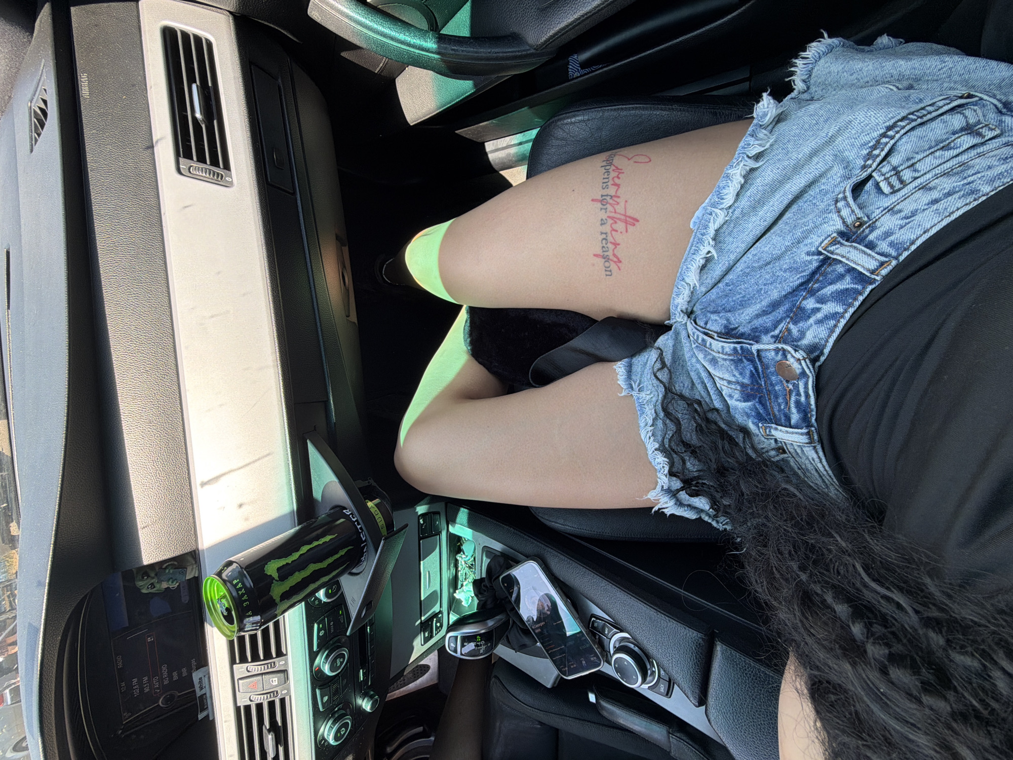 The image shows a close-up view of the interior of a car, likely in Burgas, Bulgaria. A person's legs are visible, wearing distressed denim shorts and a black top. A tattoo is visible on their thigh, with red lettering that reads "Everything happens for a reason". A can of Monster Energy drink is placed in the car's cup holder. The dashboard, gear shift, and part of the steering wheel are also visible. The overall impression is a casual, sunny day scene inside a vehicle.
