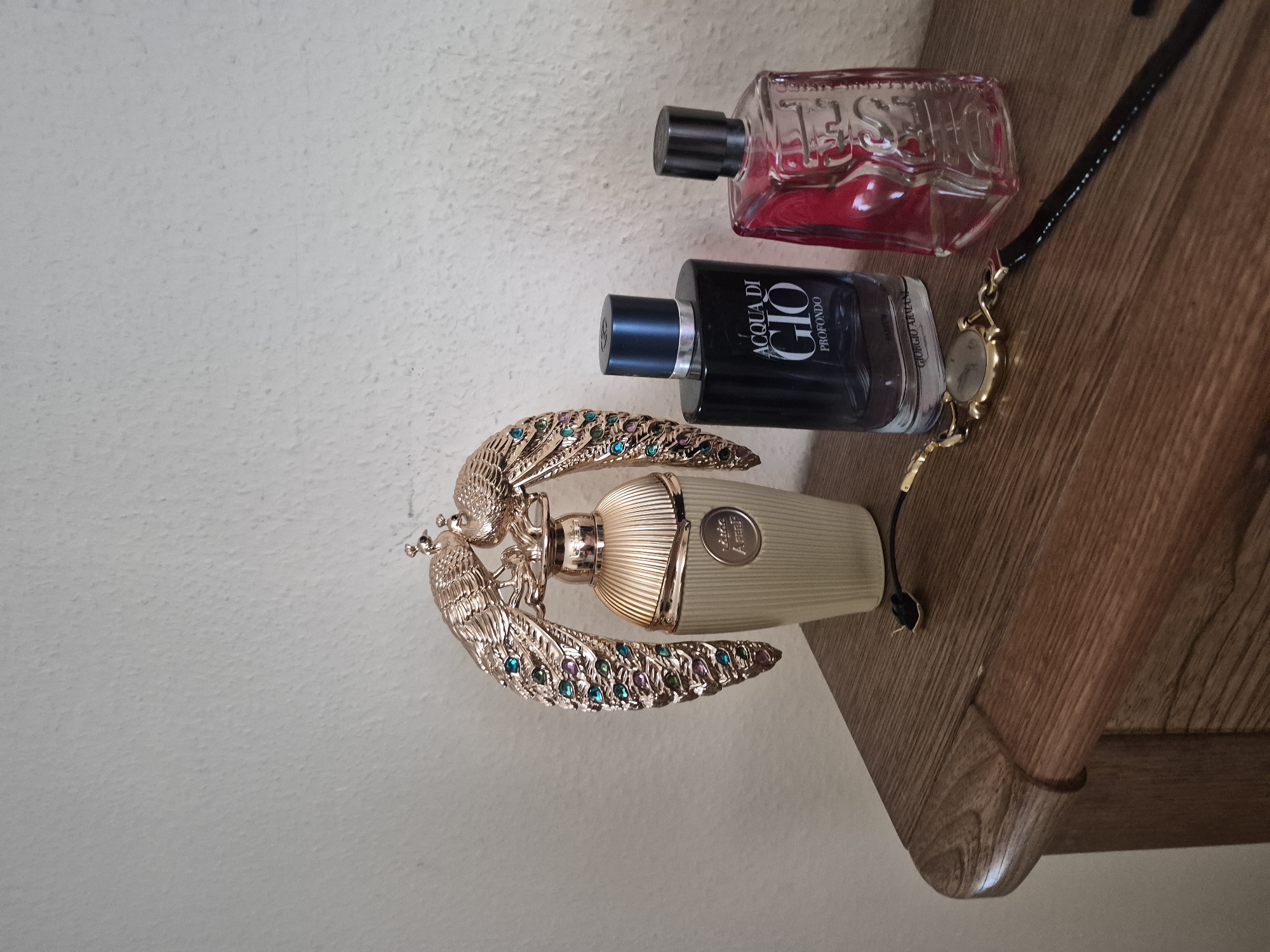 A close-up, eye-level shot shows three perfume bottles and a watch on a wooden dresser against a textured white wall. The leftmost bottle is ornate, with a gold finish and two jeweled peacock wings adorning its cap. The middle bottle is dark blue glass with a black cap, labeled "ACQUA DI GIO PROFONDO" in white text. The rightmost bottle is clear glass with a pink hue and a black cap, with faint white text. A gold watch with a black leather strap rests in front of the bottles, its face and band clearly visible. The dresser has a rounded edge and a visible wood grain. The lighting is soft and diffused, casting minimal shadows.