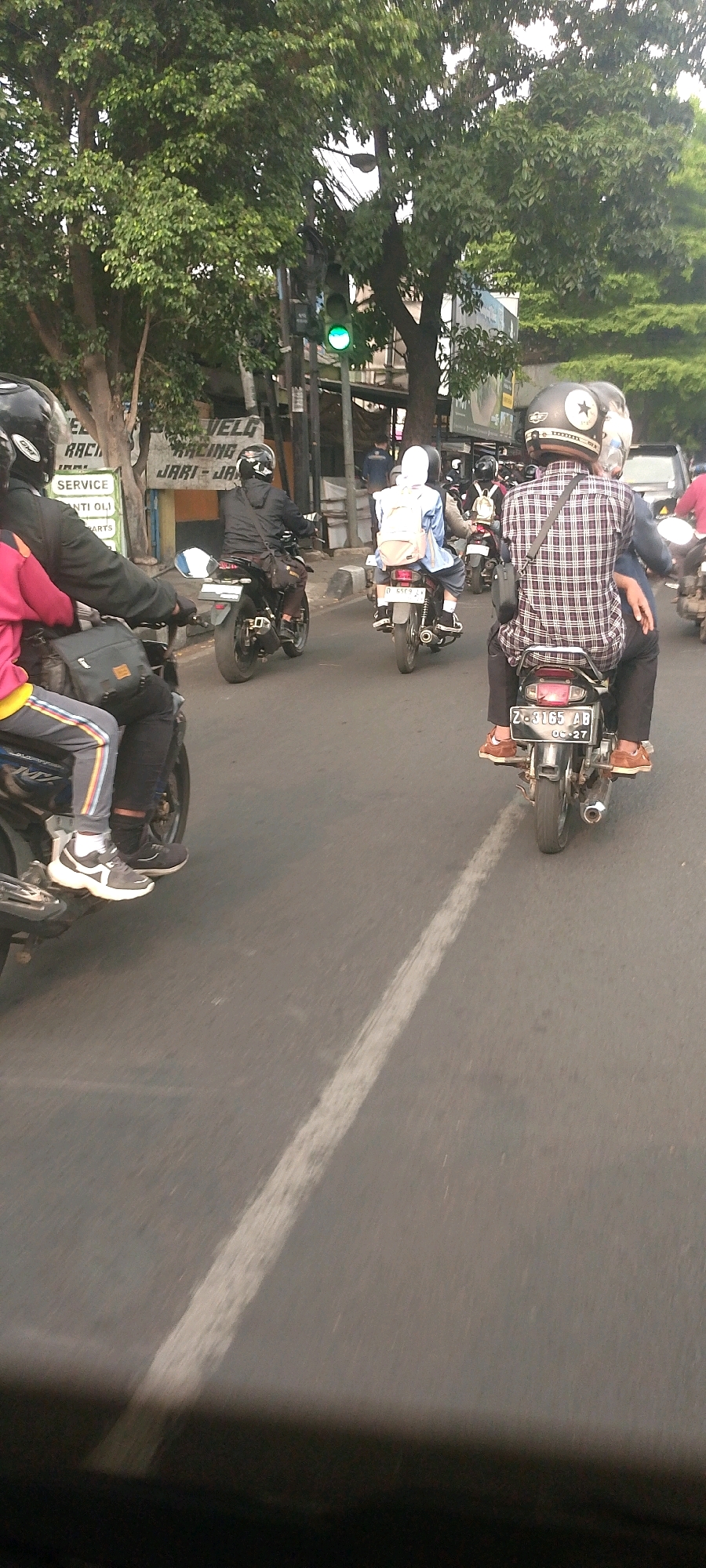 The image depicts a street scene in Bandung City, Indonesia, featuring multiple motorcycles and riders on a paved road. In the foreground, two motorcycles are visible, one on the left with a rider wearing a black jacket and a pink shirt, and another on the right with a rider in a checkered shirt. The rider on the right has a passenger behind them. The motorcycles are in motion, indicated by the blur of the road and some of the surrounding elements.
Behind the foreground motorcycles, several other motorcycles and riders are present, some also in motion. One motorcycle in the middle ground has a rider wearing a white headscarf and a backpack. Further in the background, more vehicles and trees are visible, along with a traffic light showing green. The overall environment appears to be an urban street with a mix of vehicles and foliage. The time of day seems to be daytime, with ambient light illuminating the scene. The road has a white line marking one lane.
