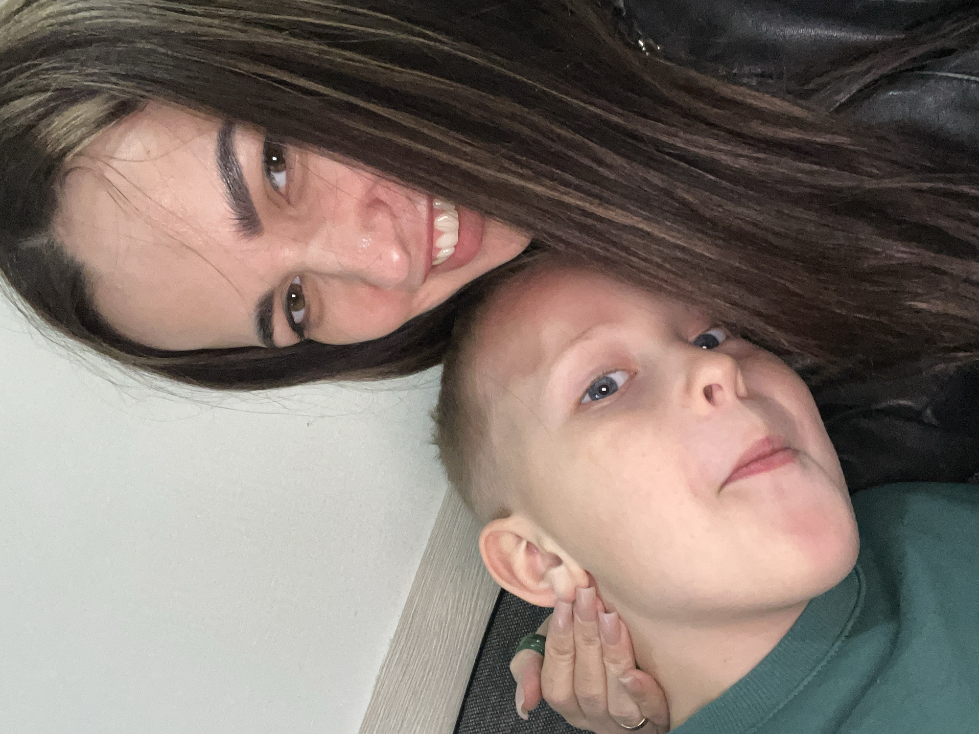 The image shows two individuals, a woman and a child, in an indoor setting. The woman has long, dark brown hair with lighter highlights, brown eyes, and is smiling, revealing her teeth. She is wearing a black leather jacket. The child has short blonde hair and blue eyes, and is wearing a green sweater. The child's face is being held gently by a hand with long, manicured nails and a green ring on one of the fingers. The background is a white wall and a gray surface. The location is Unknown, Unknown.