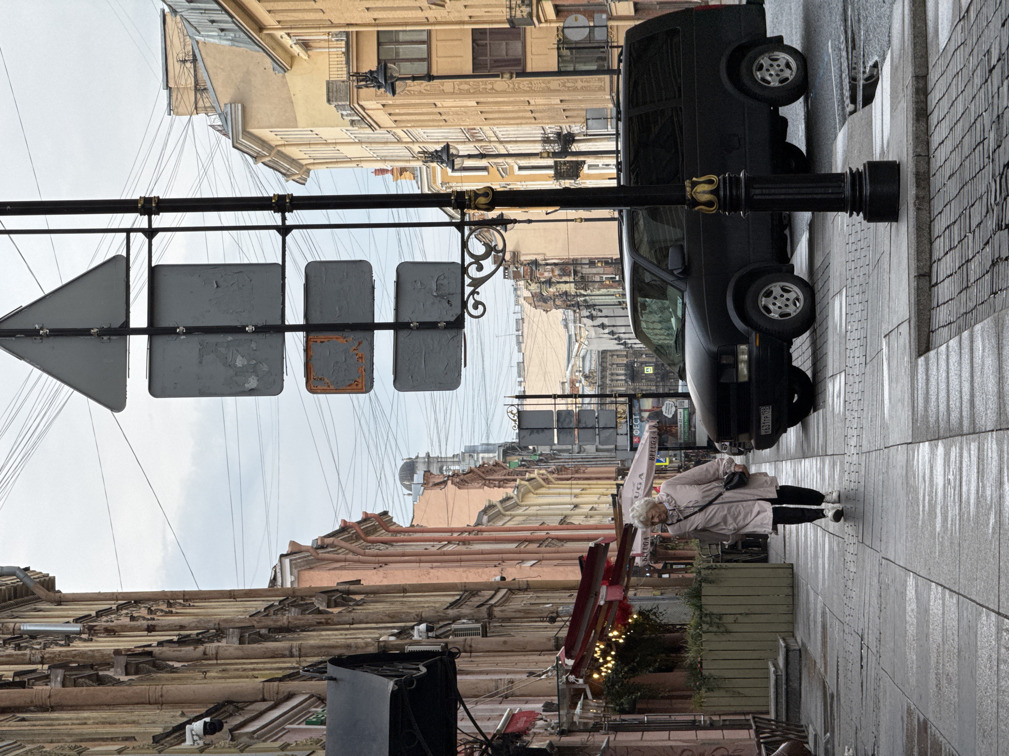 The image depicts a street in Saint Petersburg, Russia. The street is lined with old buildings of various colors, including beige, pink, and yellow. Overhead, a web of power lines crisscrosses the sky. A black van is parked on the right side of the street. A traffic sign with peeling paint is visible on the left. An elderly woman wearing a beige coat and black pants is walking on the wet sidewalk. There are plants in planters nearby, and Christmas lights are visible on a storefront. The street appears wet, suggesting recent rain.