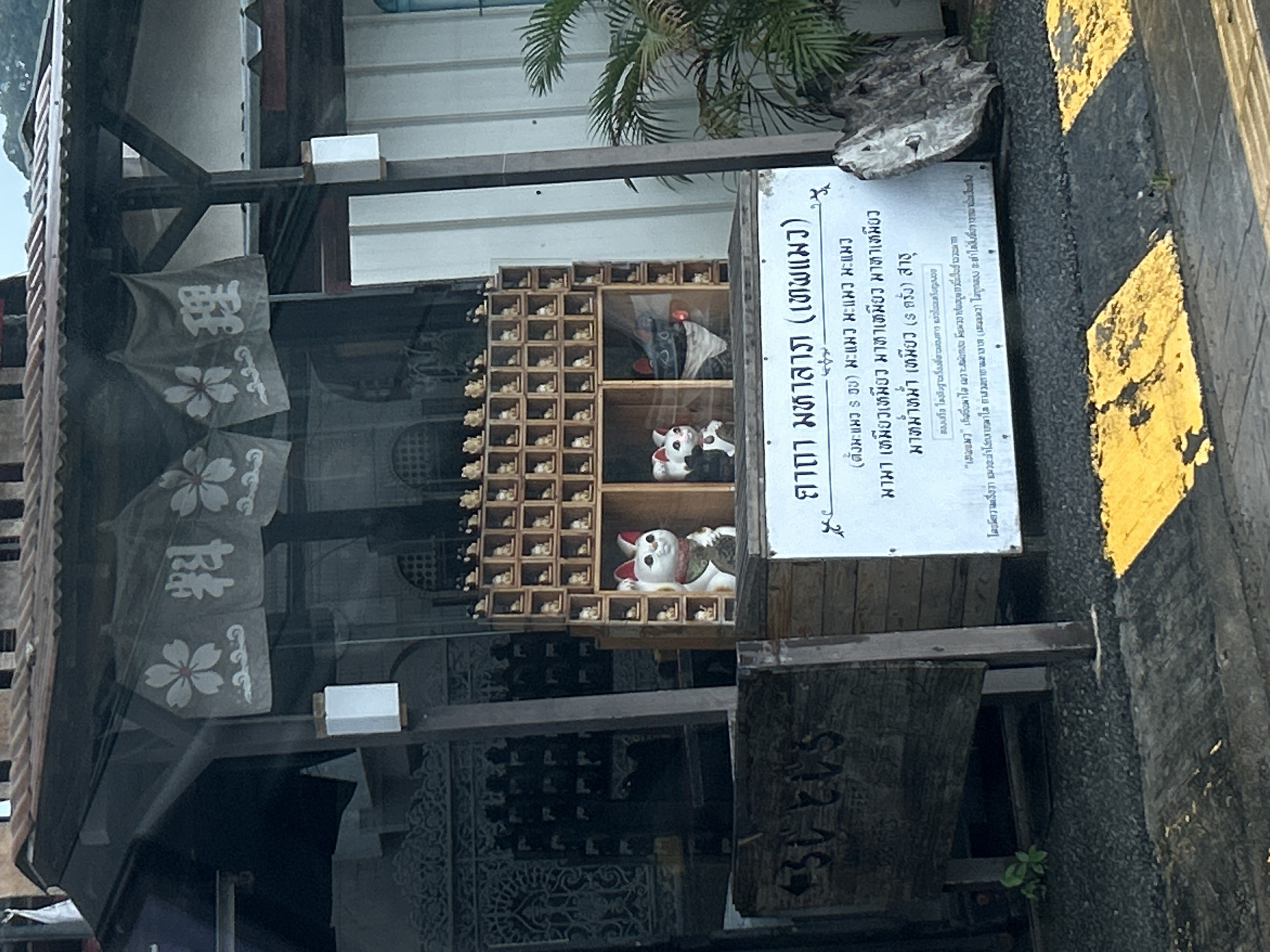 The image shows a shop with Japanese-style decorations, likely a souvenir or gift shop, in Phuket City Municipality, Thailand. In the foreground, there is a wooden sign with what appears to be Thai text. The shop itself has a traditional Japanese awning with floral and character decorations. Inside, there are numerous small figurines, possibly Maneki-neko (beckoning cats), displayed in cubbies. Two larger Maneki-neko statues are prominently featured in the front of the display. Behind the shop, a lush green palm tree is visible. The ground in front of the shop is covered with dark gravel, and there are yellow and black striped markings, resembling a pedestrian crossing or a warning zone, on the pavement. The overall scene suggests a commercial area, possibly a street or a market, in Phuket.
