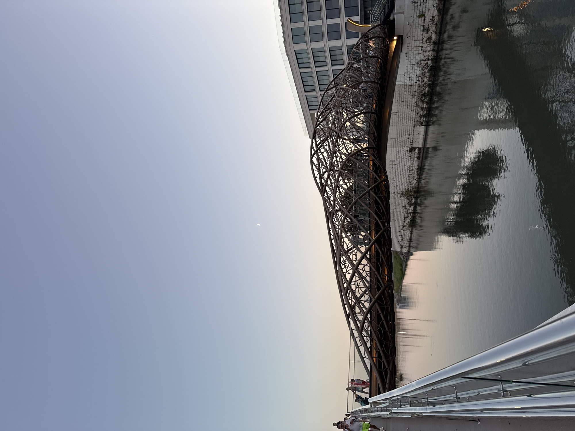 In Sochi, Russia, an ornate pedestrian bridge with a unique, interwoven metal structure spans a calm river. The bridge's design features a series of curved, interconnected metal bars creating a cage-like appearance. People are visible walking across the bridge, some silhouetted against the sky, while others are clearer in the foreground. A modern, multi-story building with a glass facade is situated on the right bank of the river, its reflection visible in the water. The sky is clear and pale blue, with a faint crescent moon visible. The overall scene suggests it is late afternoon or early evening.