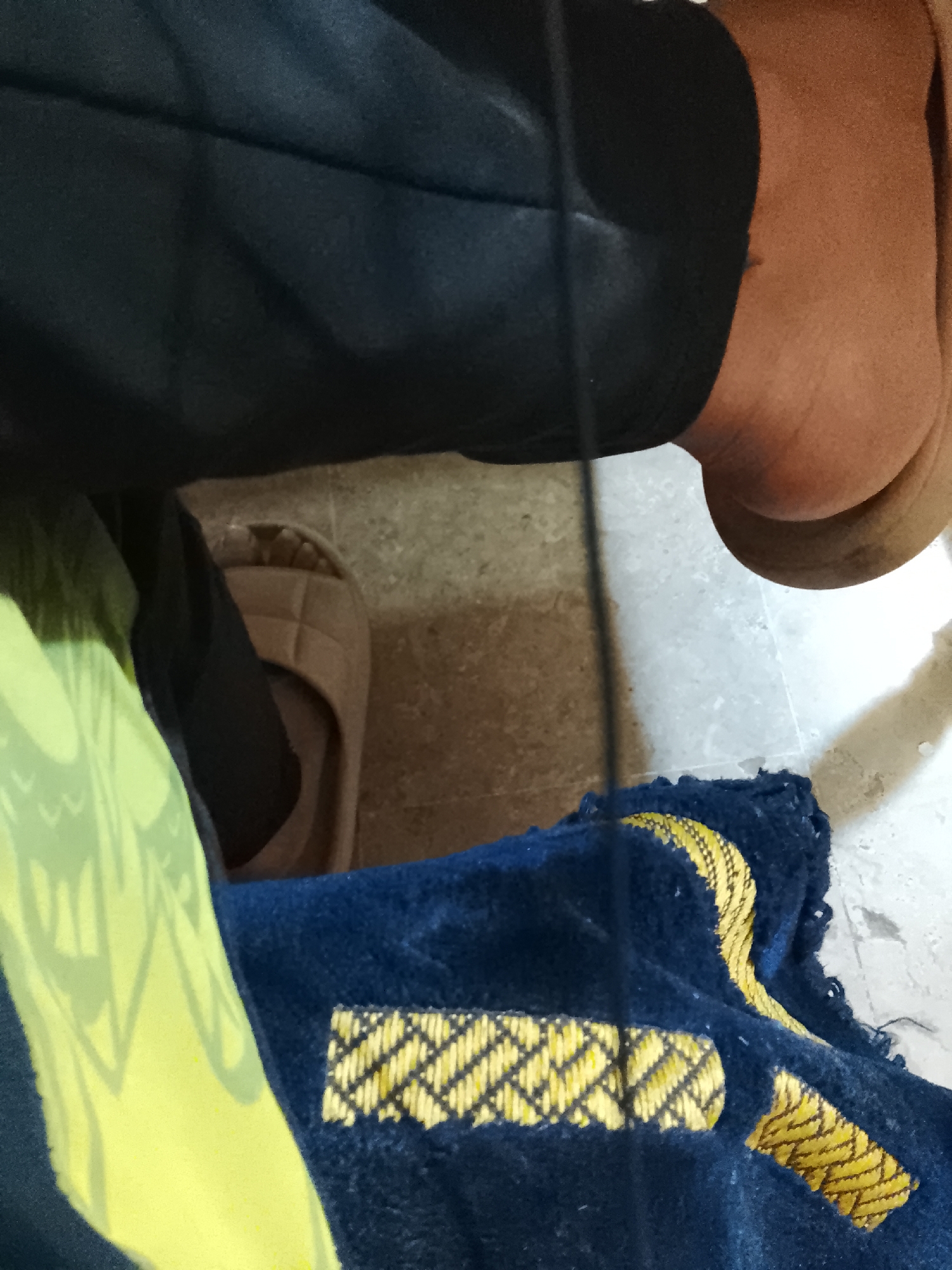 The image shows a person seated on a tiled floor. The person is wearing black pants and brown slides. A blue and yellow rug is visible next to the person. It is not possible to confirm this is in Multan, Pakistan.