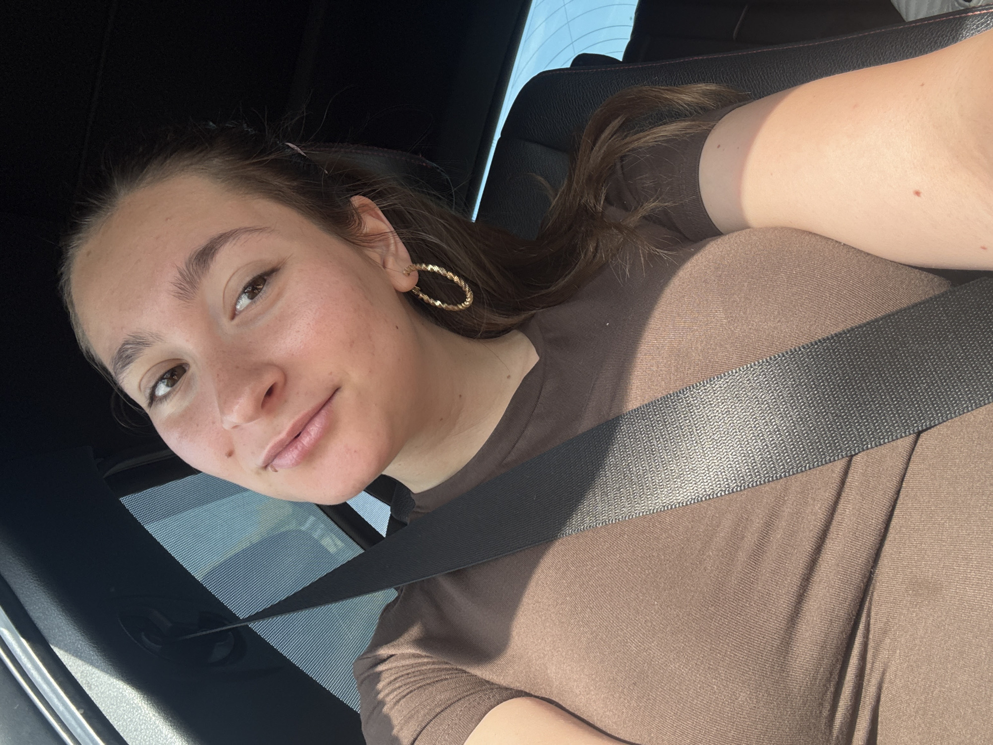 The image shows a woman sitting in the driver's seat of a car, wearing a brown t-shirt and a seatbelt. She is looking at the camera and smiling slightly. The interior of the car is visible, including the dashboard and the side window with a sunshade. The woman has long brown hair and is wearing gold hoop earrings. The lighting suggests it is daytime, with sunlight coming through the window. There are no other people or objects of note in the immediate vicinity. The scene is a typical interior of a vehicle.