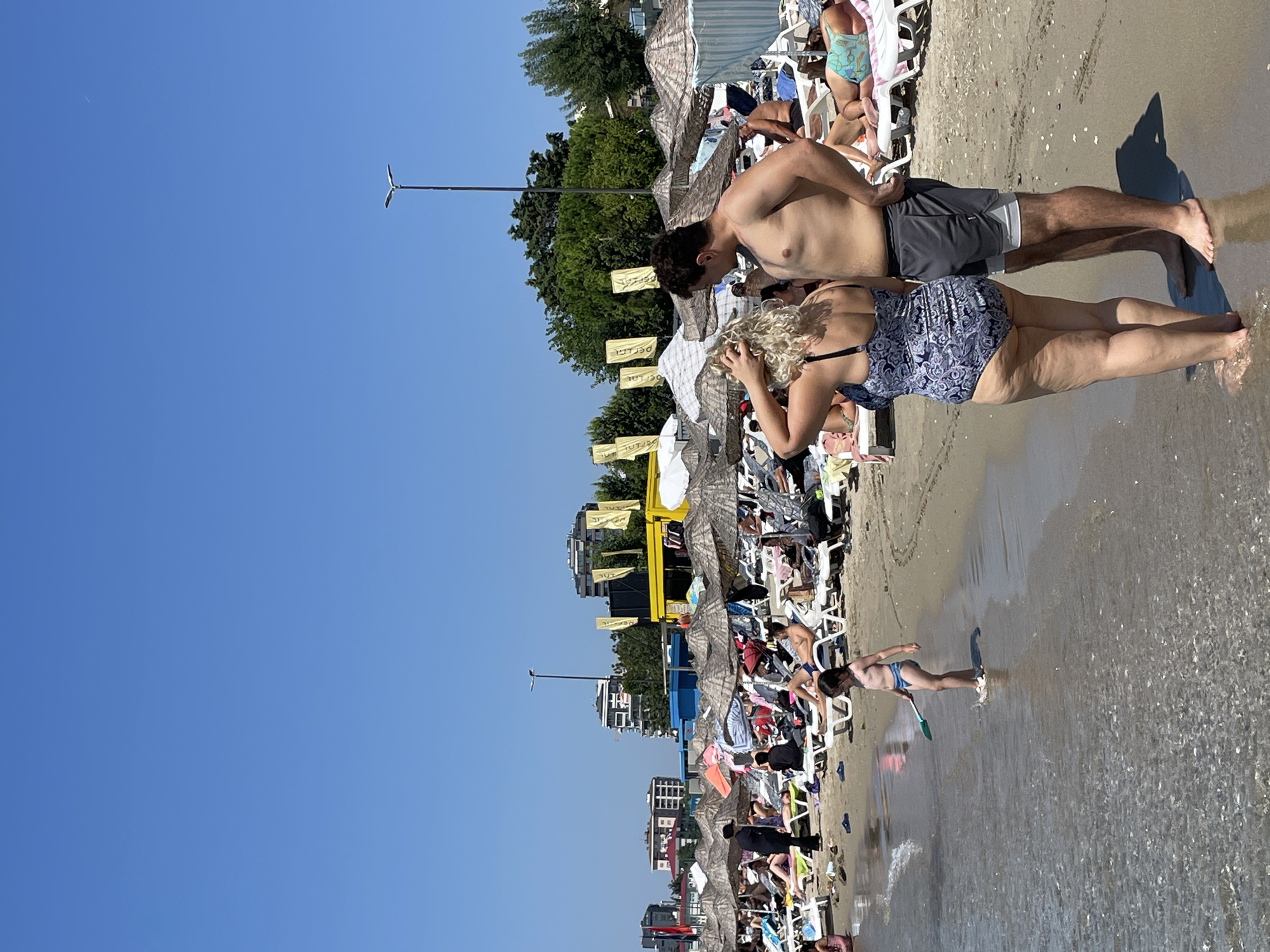 The image depicts a crowded beach scene on a sunny day, with many people relaxing under umbrellas and on lounge chairs. In the foreground, a couple stands facing away from the camera, with the woman wearing a patterned swimsuit and the man wearing swim trunks. To the left, a young child in a swimsuit is playing in the shallow water with a blue shovel. The background shows more beachgoers, a row of umbrellas and lounge chairs, and buildings and trees in the distance. The presence of flags and buildings suggests an urban or semi-urban beach location. This scene could be representative of a beach along the coast of Kadıköy, Turkey, a district known for its beaches and vibrant atmosphere.