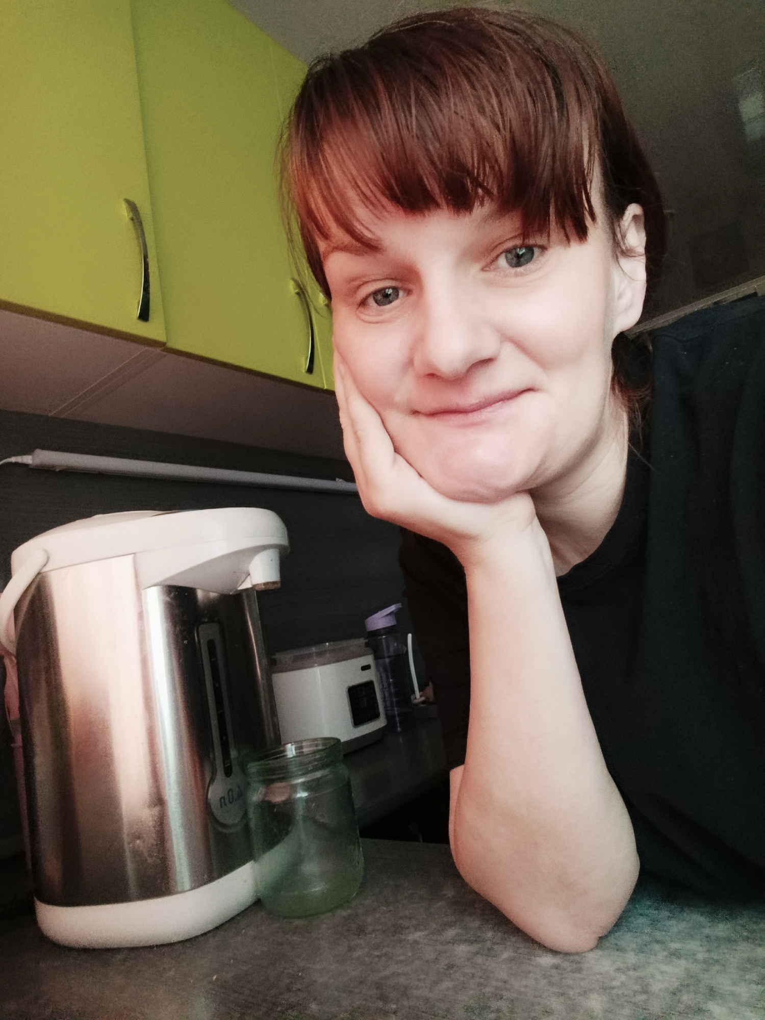 The image shows a person with short, reddish-brown hair and bangs, resting their face on their hand. They are wearing a black shirt. Behind them is a kitchen counter with a stainless steel water boiler and a glass jar. Above the counter are lime green cabinets. An electrical outlet strip is visible on the wall behind the boiler. The location is Unknown, Unknown.