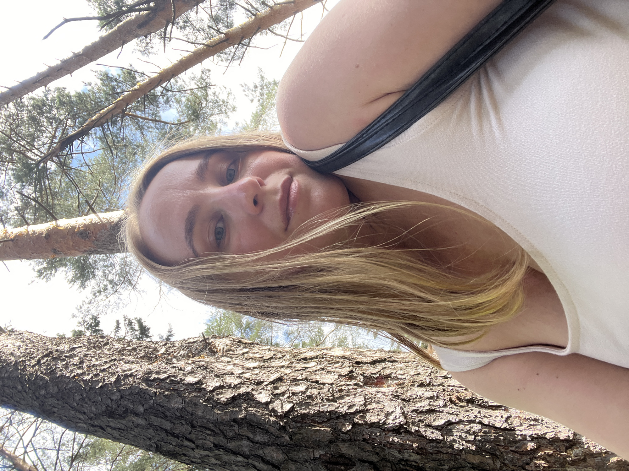 A blonde woman is taking a selfie outdoors. She is wearing a light-colored tank top and has a black strap from a bag across her shoulder. She is positioned to the right of the frame, with the trunk of a large tree occupying the left side. The background consists of more trees and a bright sky, suggesting a wooded area. The image does not provide any information about the city of Chaikovsky, Russia, or any specific location within it.