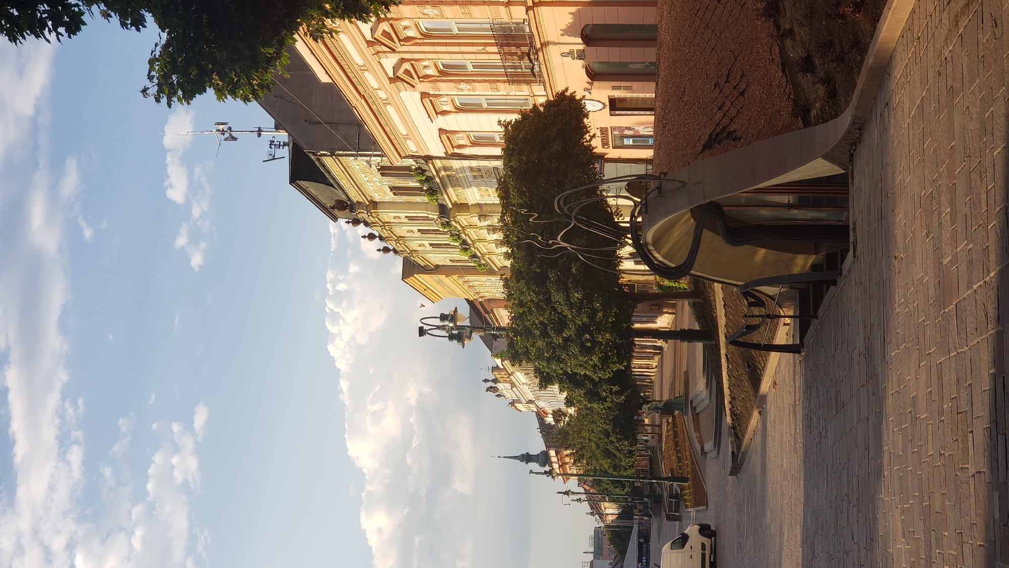 The image depicts a street in the city of Prešov, Slovakia, likely on Hlavná ulica (Main Street) given the architecture and urban setting. The scene is captured during the daytime, with soft, warm sunlight casting long shadows, suggesting it is either early morning or late afternoon.
The left side of the street features a cobblestone pavement and a sidewalk lined with trees. A white van is parked on the left side of the frame. Further down the street, a statue of a figure is visible amongst the trees, and a spire of a church or tower is prominent in the distance. Several streetlights with multiple lamps line the sidewalk.
On the right side of the street, a row of historic buildings with ornate facades and balconies are present. These buildings appear to be commercial establishments, with shop windows visible. In the foreground on the right, a modern, sculptural entrance with a wavy metal design is situated at ground level, leading down to what appears to be an underground space. The area around this entrance is landscaped with brown mulch and some small plants.
The sky is partly cloudy, with a mix of blue sky and white, fluffy clouds. The overall atmosphere is calm and picturesque, showcasing a blend of historical architecture and contemporary design within an urban environment. There are no people visibly present in the image.