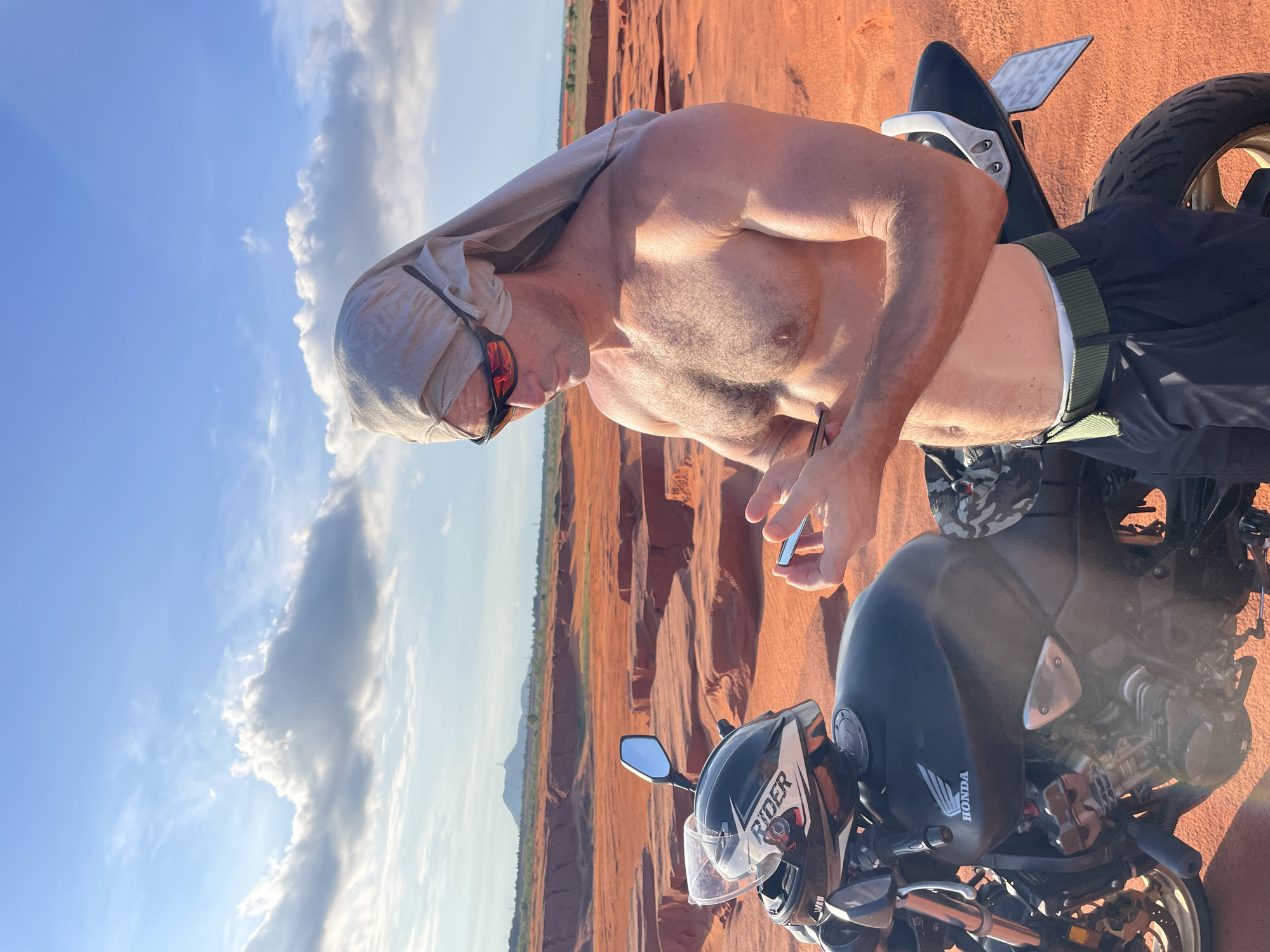 In the arid landscape of Mui Ne, Vietnam, a man stands next to a black Honda motorcycle. He is shirtless, wearing black pants and a green belt, with sunglasses on and a light-colored cloth draped over his head. His hands are occupied with a small electronic device, possibly a phone. The environment is characterized by striking red sand dunes with layered geological formations, under a bright blue sky dotted with white clouds. In the distance, a lone mountain peak rises from the horizon. The motorcycle is equipped with a black helmet displaying "RIDER" text and a transparent visor. The scene conveys a sense of adventure and exploration in a remote, scenic setting.