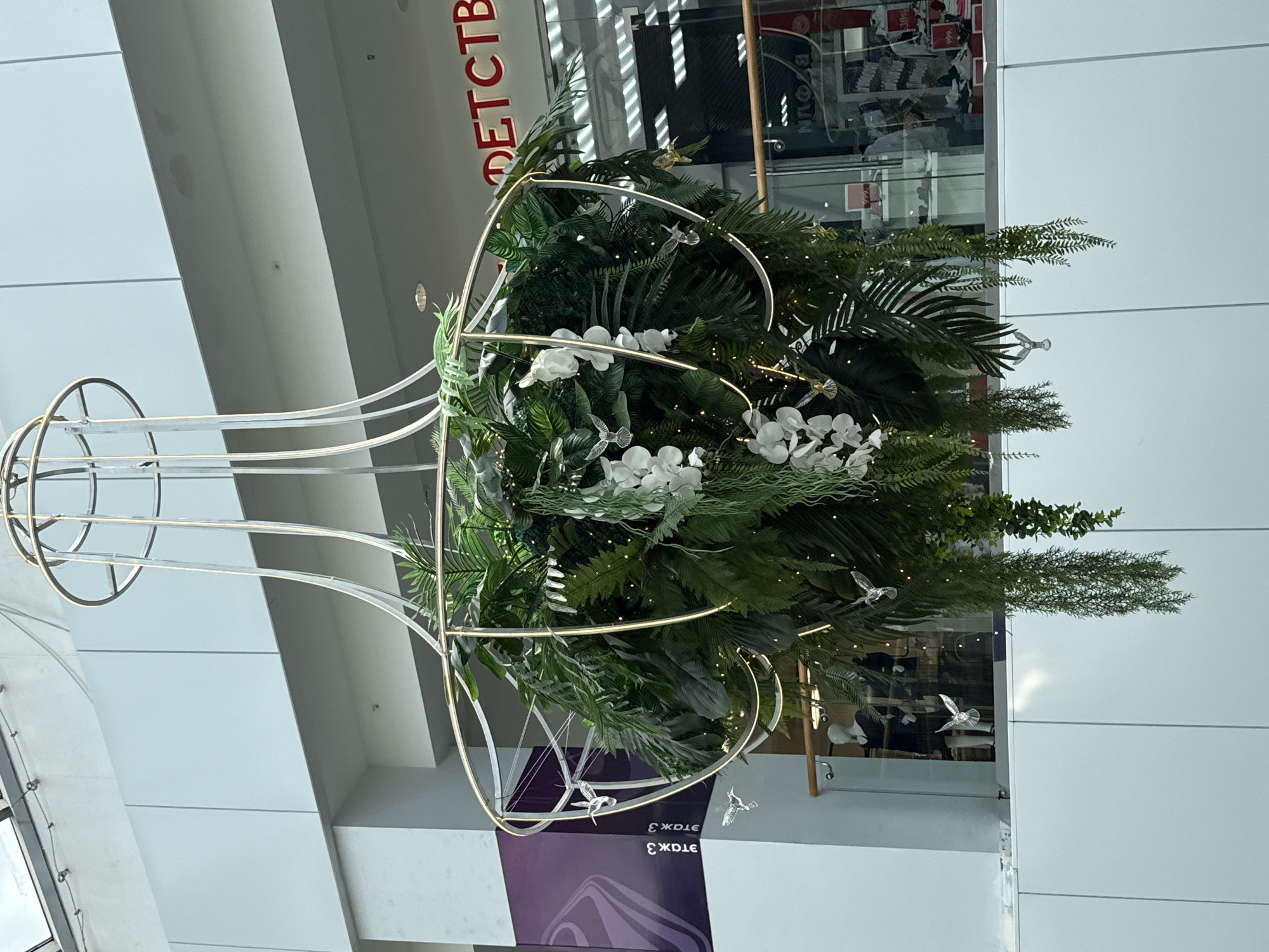 The image shows a decorative hanging installation in what appears to be a shopping mall. The installation is a large, globe-like structure filled with artificial green foliage and white artificial orchids. It is suspended from the ceiling by thin, metallic rods and has a golden-colored hoop around its perimeter. Several small, transparent hummingbird figurines are attached to the structure, appearing to be in flight. Small, warm-toned lights are also interspersed within the foliage. In the background, there are store windows with mannequins visible, and a sign with Cyrillic text that includes "ДЕТСТВ" (likely part of "ДЕТСТВО" meaning "childhood" or "children's store") is visible. Another sign shows "ЭТАЖ 3" (Floor 3). The overall environment suggests an indoor commercial space.