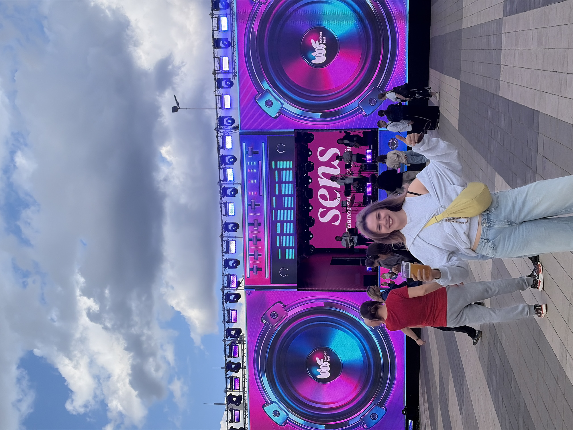 This image captures a daytime outdoor event, likely a music festival, under a partly cloudy sky. The focal point in the foreground is a young woman with light purple hair, smiling and giving a thumbs-up while holding a plastic cup of beer. She is wearing a light gray off-the-shoulder sweatshirt, light blue jeans, and a yellow crossbody bag. Behind her, a large LED screen displays a vibrant graphic of a speaker with swirling pink and blue lights and the text "sens". The screen is flanked by smaller graphics and lighting rigs. A stage with a band performing is visible behind the LED screen. Other attendees can be seen milling about in the background. The ground is paved with interlocking gray and black rectangular stones. The overall atmosphere suggests a lively and festive gathering. The text "belbet fest" is visible on the speaker graphic, indicating the name of the event. The presence of the "belbet fest" logo suggests this event is taking place in Minsk, Belarus, as Belbet is a Belarusian betting company.