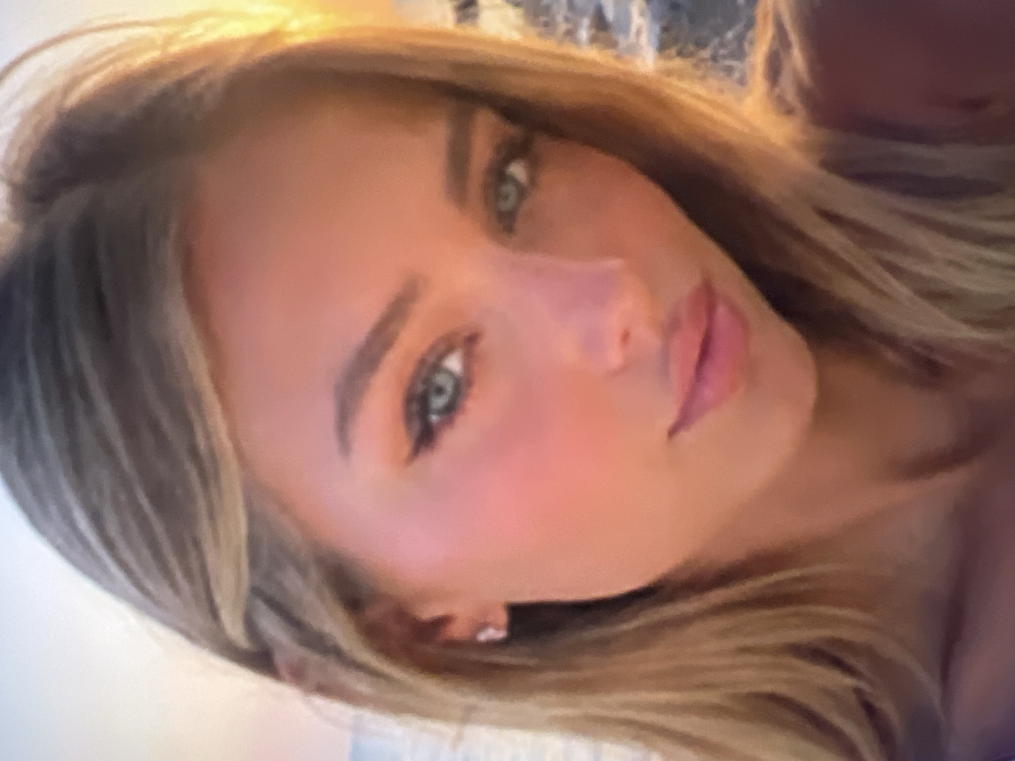 This is a close-up, head-and-shoulders shot of a young woman with blonde hair. Her head is tilted, and she is looking off to the side. Her eyes are light blue and her lips are slightly parted. She is wearing light makeup, including eyeliner and blush. The lighting suggests it might be daytime, possibly with natural light from a window or outdoors. There are no visible objects or text in the image. The background is blurred and indistinct, making it difficult to determine the setting. The woman's expression is neutral or slightly contemplative. The image appears to be a selfie.