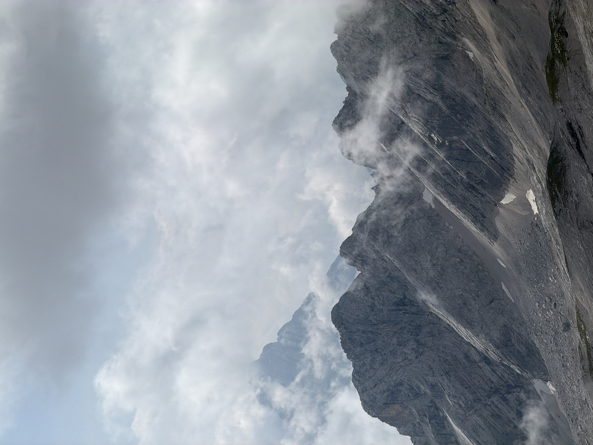 The image depicts a dramatic mountain landscape with jagged peaks piercing through swirling clouds. The foreground features rugged, rocky terrain with visible stratification and patches of snow. The middle ground showcases several prominent mountain ridges, their sharp contours softened by mist and low-hanging clouds. In the background, more distant mountain peaks are shrouded in fog, creating a sense of depth and mystery. The sky above is overcast, with heavy, grey clouds dominating the upper portion of the frame, interspersed with lighter, wispy formations. The overall impression is one of a wild, untamed alpine environment. The specific location in Wildermieming, Austria, is not explicitly identifiable from the image alone, but the scene is characteristic of the Ötztal Alps region. There are no people, animals, or man-made structures visible in the frame.