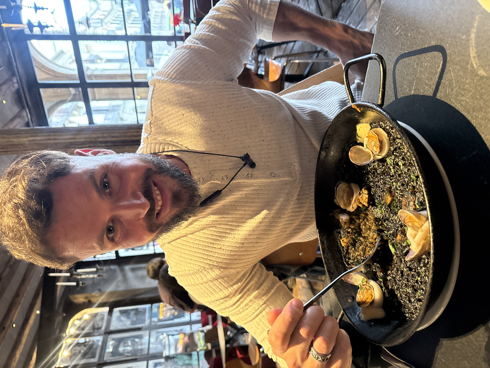 A man sits indoors at a restaurant in Barcelona, Spain, holding a pan of black rice paella with clams and wielding a spoon, poised to eat. He is wearing a white, textured shirt with a visible headset around his neck and a ring on his finger. The pan sits on a black surface. In the background are a window with multiple panes and other restaurant patrons.