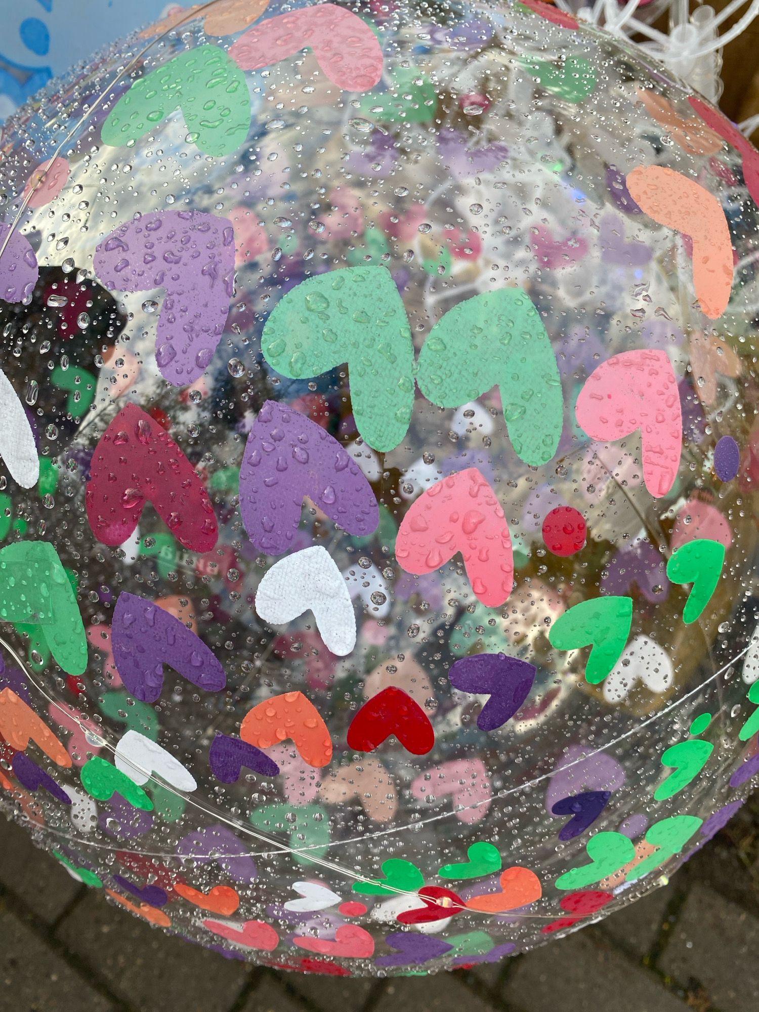 The image displays a close-up of a clear, inflatable ball filled with colorful heart-shaped confetti. The ball is covered in numerous small water droplets, suggesting it is outdoors and has recently been exposed to rain. The heart confetti is varied in color, including pink, purple, green, white, red, and orange. The background is blurred, but appears to be an outdoor setting, possibly a street or paved area with some greenery visible in the distance. There are no people or discernible actions taking place in the image. The location is Ivanteyevka, Russia, though no specific landmarks or contextual clues within the image confirm this location.