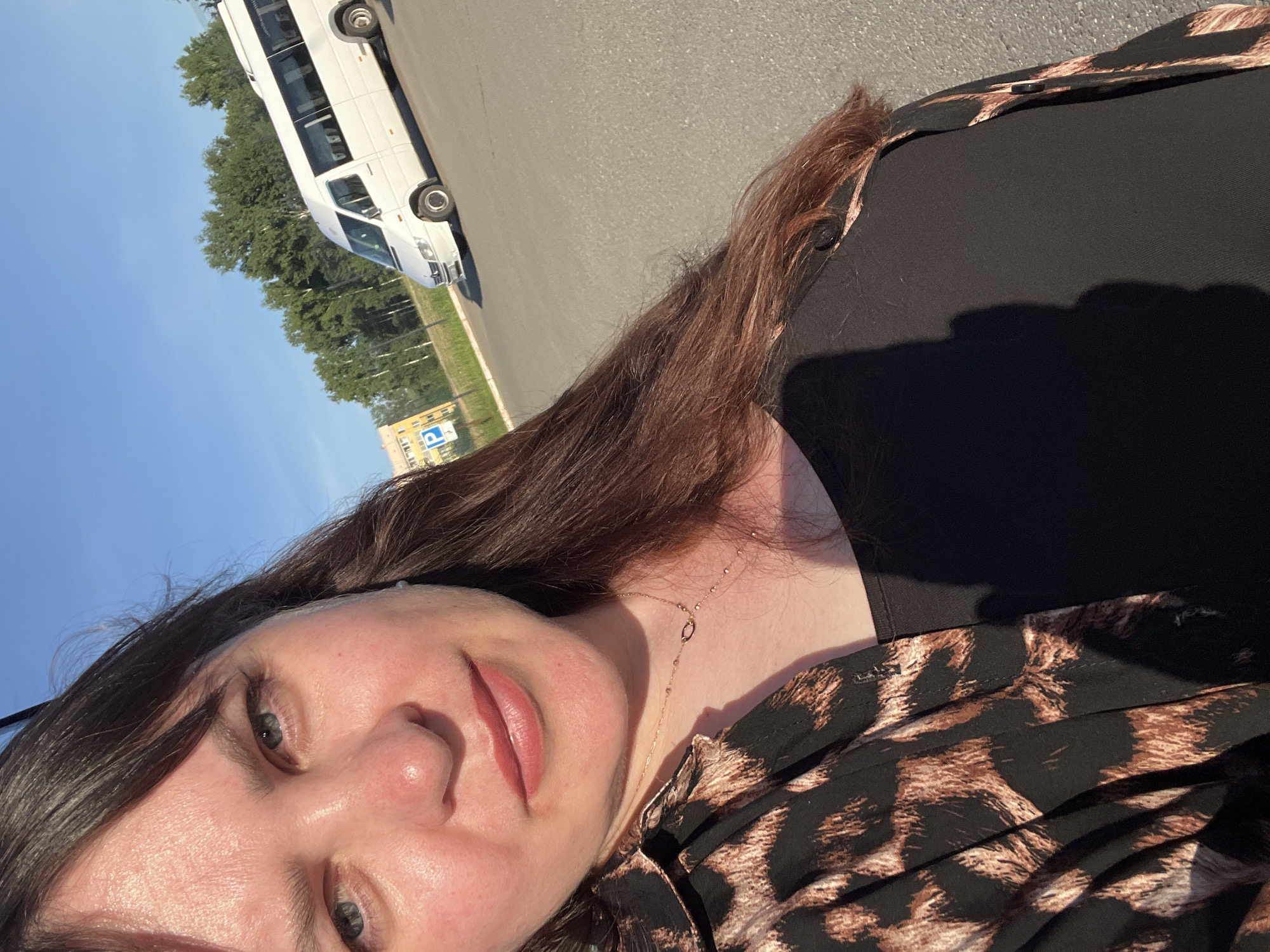The media file is a portrait of a person in an outdoor setting. The subject is a woman with long, dark brown hair, fair skin, and blue eyes. She is wearing a black top with a leopard print overlay and a delicate gold necklace. Her makeup includes pink lipstick and subtle eye makeup. The image is taken from a slightly high angle, focusing on her face and upper body.
In the background, a white mini-bus is parked on an asphalt surface. Beyond the bus, there are green trees and a clear blue sky, suggesting it is daytime with good weather. A yellow building with a blue parking sign is visible in the distance, providing a hint of the location. The overall lighting suggests it is either morning or late afternoon, given the angle of the shadows.
The subject's facial expression appears relaxed and natural, with a slight smile. The composition is a selfie, indicated by the framing and the perspective. The scene appears to be a parking lot or roadside area, possibly near a residential or commercial zone in Noviy, Russia. The overall impression is a casual snapshot taken during a bright, sunny day.
