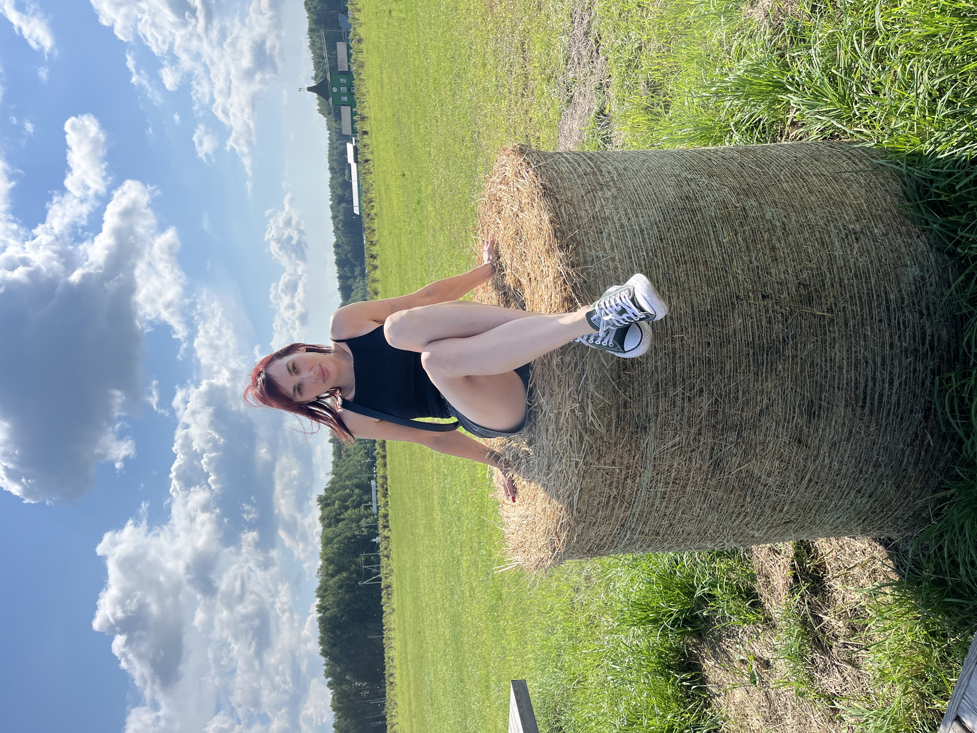In Akhlebaikha (Kovriginskii S/S), Russia, a young woman with red hair is sitting on a large, round bale of hay in a grassy field under a bright blue sky with fluffy white clouds. She is wearing a black tank top and dark shorts, with black and white Converse sneakers on her feet. Her legs are crossed, and her left hand rests on the hay bale. In the background, a line of green trees stretches across the horizon, and a distinct green building with a pointed roof and weather vane is visible. The scene appears to be a rural setting on a sunny day.