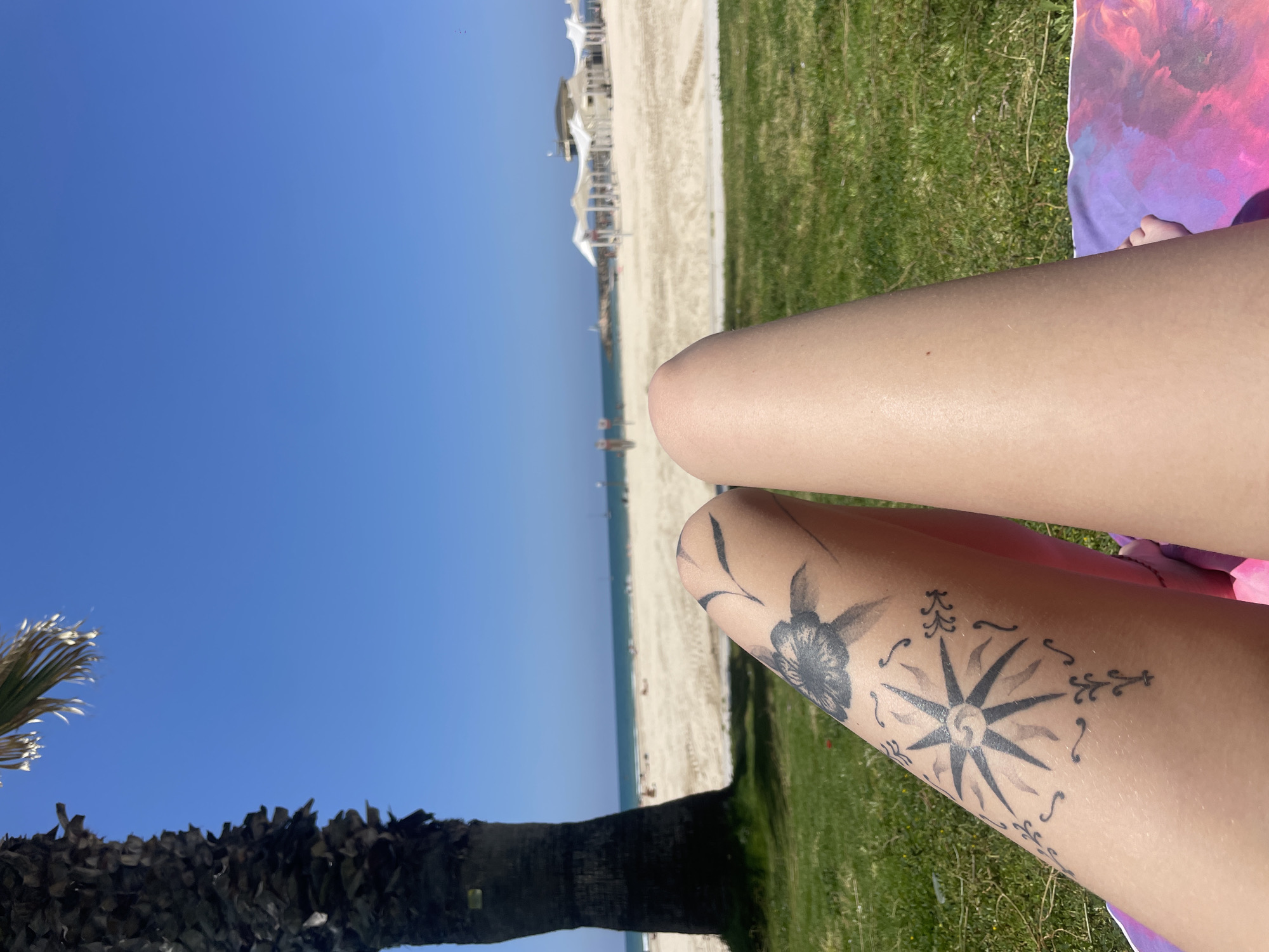 The image shows a pair of legs against a backdrop of a beach and ocean. One leg has a tattoo featuring floral and compass designs. The scene includes a beach with white sand, an ocean, clear blue sky, palm tree, grass, beach establishments, a partially visible colorful blanket, and people in the distance. This image was taken in Herzliya, Israel.