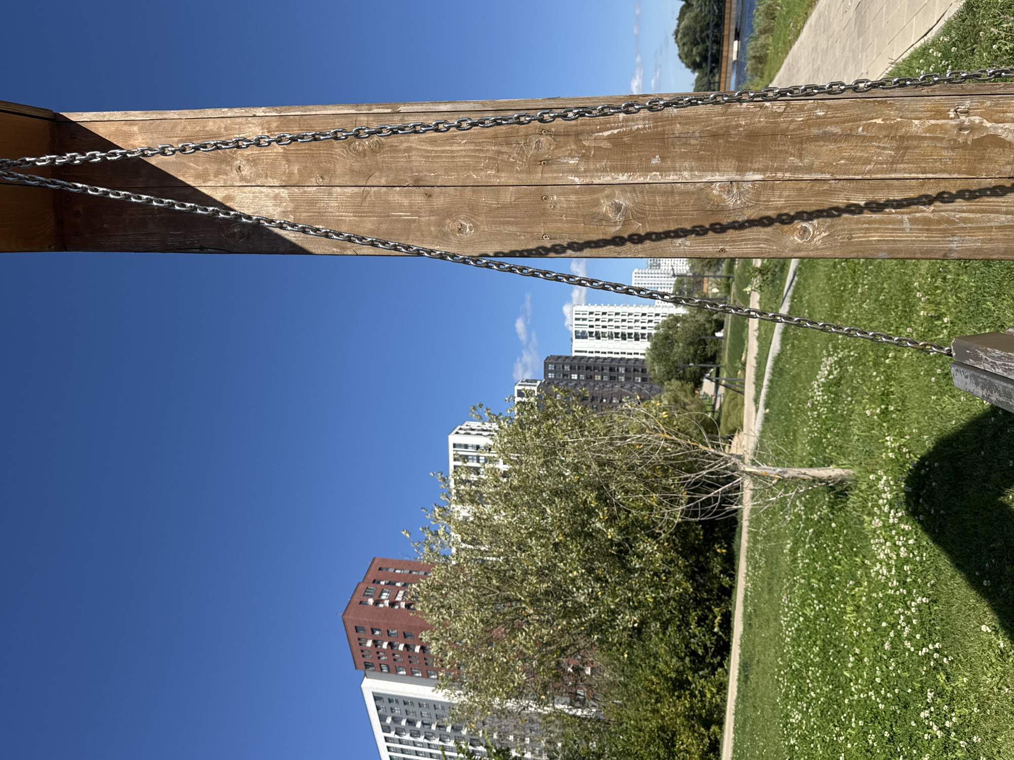 The image depicts a sunny day in a park in Kommunarka, Russia, with a clear blue sky overhead. In the foreground, a wooden post stands to the right, adorned with two thick metal chains. The post is bathed in sunlight on the right side, while the left side is cast in shadow. To the left of the post, a lush green lawn is visible, dotted with small white flowers. A small tree with sparse foliage stands on the lawn, casting a faint shadow. In the middle ground, a line of trees and bushes forms a verdant backdrop. Beyond the trees, several modern apartment buildings are visible, characterized by their white, grey, and red facades. One building in particular, with a striking red facade, stands out. Further in the distance, a body of water can be seen, with a bridge crossing over it. The overall scene suggests a well-maintained urban park, likely a recreational area for residents of the surrounding apartment complexes. The absence of people indicates a quiet, peaceful moment captured in the photograph.