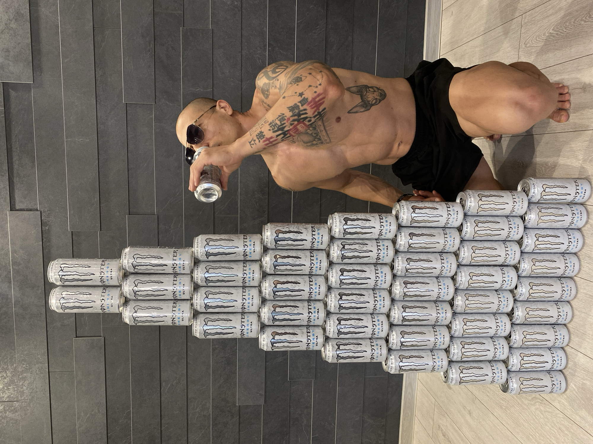 A muscular, bald man with sunglasses and numerous tattoos is kneeling indoors on a gray and beige tiled floor. He is shirtless, wearing black shorts, and drinking from a can of Monster Energy Ultra drink. In front of him, a pyramid of Monster Energy Ultra cans is stacked, forming an imposing structure. The man appears to be the central figure of the image. The location is an indoor space. The image does not provide enough information to determine whether it is in the city of Pleven, Bulgaria.