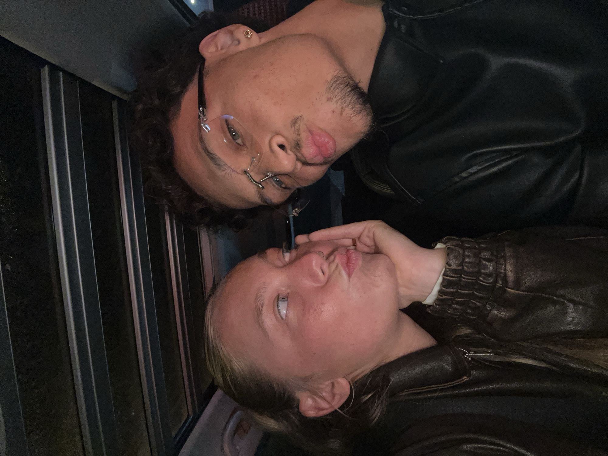 Two individuals are captured in an off-kilter selfie. The person on the left has fair skin, light blonde hair, and blue eyes. They are wearing a brown leather jacket and puckering their lips towards the camera. Their hand is also raised near their face. The individual on the right is a male with darker skin, curly dark hair, and glasses. He has some facial hair and is also puckering his lips. He is wearing a black leather jacket. The background consists of vertical metal slats, suggesting a window or enclosure. The location and city information of Noisy-le-Grand, France, cannot be definitively determined from the visual elements in the provided image.