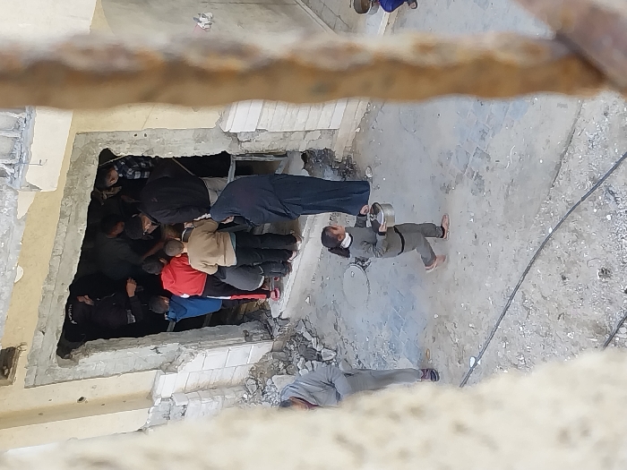 The image depicts a scene in Khan Yunis, Palestinian Territory, observed from an elevated perspective, showing a group of people gathered around a damaged building entrance and individuals moving across a debris-strewn ground.
At the center, a large, rectangular opening in a light-colored building exhibits significant structural damage, with ragged edges of concrete and exposed rebar. A crowd, consisting of approximately ten to fifteen individuals, predominantly men and boys of varying ages, is clustered within and directly outside this darkened doorway. Their clothing includes jackets and sweaters in colors such as red, blue, and tan. One woman, dressed in a black abaya or long dress, stands to the right of the crowd near the entrance. A partially visible individual inside the doorway wears a dark top with white text, possibly "POLICE," on the back.
In the foreground, a young girl, estimated to be 7-10 years old, walks from left to right. She is dressed in a grey tracksuit with black trim and carries a lidded silver metal pot in her left hand. To her immediate left, another individual in grey attire is partially visible. On the far right, a small portion of a person, possibly a child, is visible crouching or seated, holding another pot.
The ground surface is uneven, composed of dirt, cracked concrete, and scattered rubble. A circular manhole cover is present in the middle ground, and black cables traverse the area. Blurred architectural elements frame the scene on both the left and right sides. The building structure itself shows signs of damage, with debris around the entrance. The overall scene conveys an environment impacted by damage, with people engaged in activities possibly related to aid distribution or resource collection.