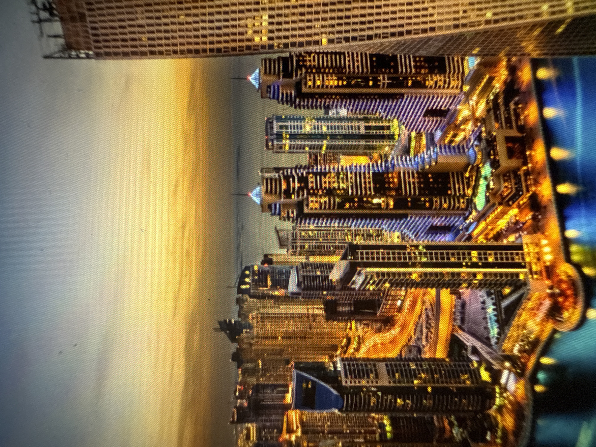 The image displays a sprawling cityscape at twilight or night, located in the city of Blagoevgrad, Bulgaria. Numerous illuminated skyscrapers of varying heights dominate the scene, featuring a blend of modern architectural designs. One prominent, very tall building is partially visible on the far right, extending vertically out of the frame. Many structures exhibit intricate patterns of illuminated windows in yellow, white, and blue tones. A wide body of water, possibly a canal or bay, occupies the immediate foreground, reflecting scattered lights from the adjacent buildings and infrastructure. Intersecting roadways and elevated highways are visible, marked by bright trails of orange and yellow light, indicating vehicular movement. The sky transitions from a light blue hue at the top to a golden-orange glow at the horizon, suggesting either dawn or dusk. No specific individuals or discernible actions are present within the frame.