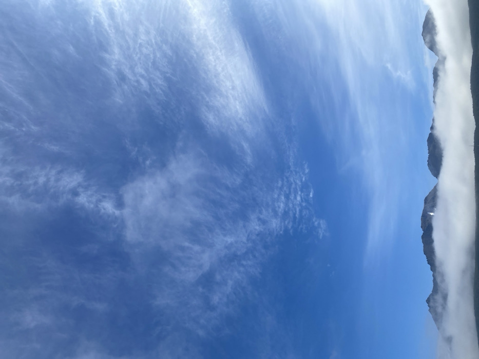 This image depicts a mountainous landscape in Ushuaia, Argentina, under a vast blue sky with scattered cirrus clouds. The upper two-thirds of the frame are dominated by a clear azure sky, transitioning to lighter blue towards the horizon, marked by wispy white cloud formations. The lower third of the image features a dark, rugged mountain range with multiple peaks, partially obscured by a dense, horizontal band of white fog or low-lying clouds that covers their bases and lower slopes. A single, distinct patch of bright white, likely snow or ice, is visible on one of the central mountain peaks, reflecting light. No people, animals, or artificial structures are discernible within the frame.