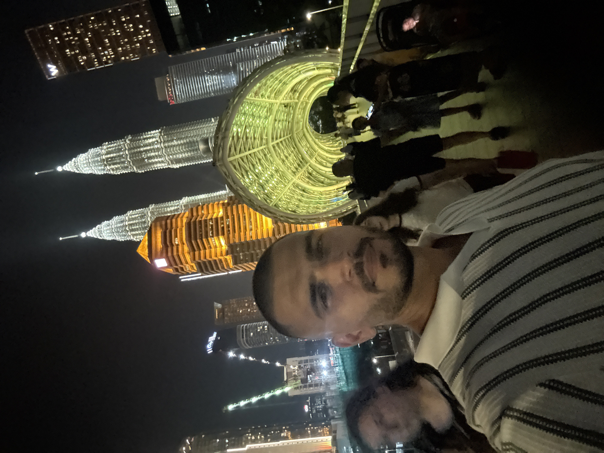 The image shows a man and a woman in front of the Petronas Towers in Kuala Lumpur, Malaysia, at night. The man is wearing a striped shirt and has a beard. The woman is in the foreground and her face is out of focus. Behind them is a pedestrian bridge lit with green and yellow lights. Several other people are walking through the bridge. In the background are several skyscrapers lit up at night.