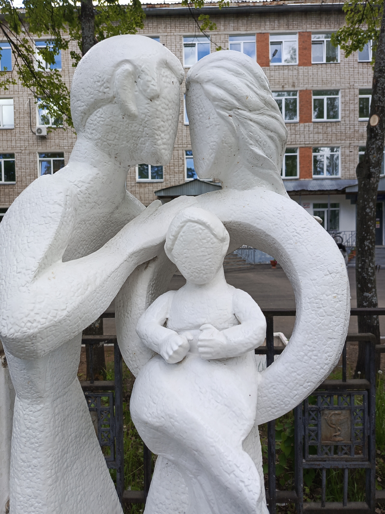 The image shows a white concrete sculpture of a family, consisting of a man, woman, and child, located in the city of Chaikovsky, Russia. The sculpture is positioned in front of a three-story brick building with white-framed windows and red brick accents. The man and woman are depicted with their heads close together, appearing to be kissing. The woman has her arm around the child, who is sitting in front of her. A black metal fence is partially visible behind the sculpture, adorned with a design featuring a snake wrapped around a staff, suggesting the presence of a medical facility. Green trees and foliage are also visible in the background.