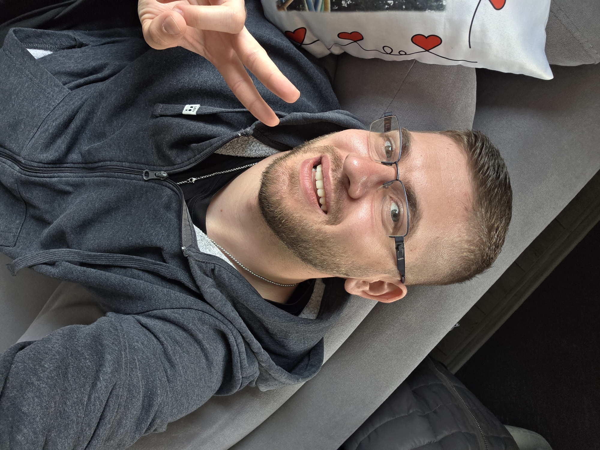 A man is lying on a gray couch, wearing a gray zip-up hoodie, a black t-shirt, and glasses. He is making a peace sign with his right hand and smiling. A white pillow with red heart designs is next to him. The location is not discernible from the image.