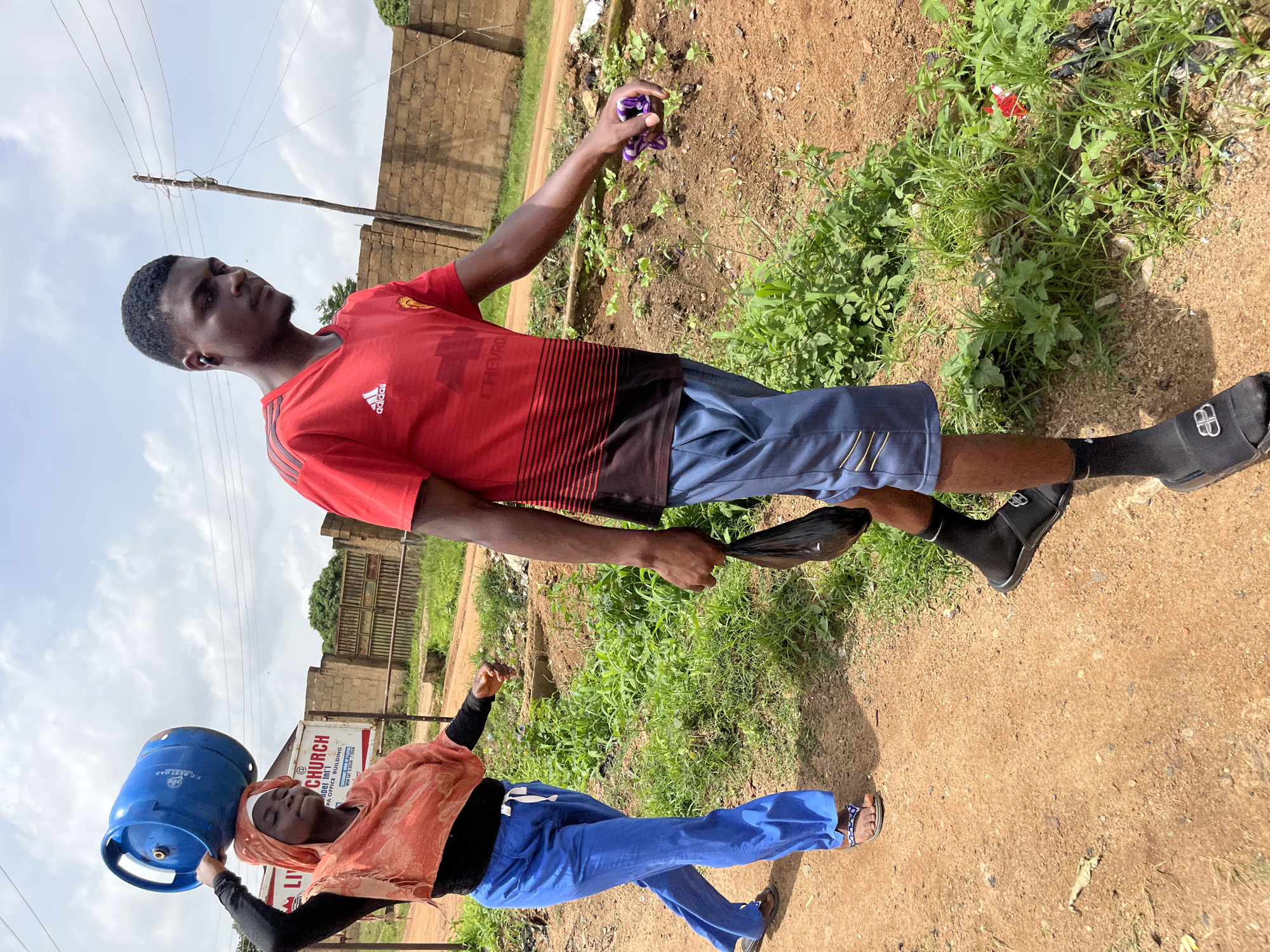 In the city of Bukuru, Nigeria, a young woman and a young man walk along a dirt path. The woman, wearing blue pants and an orange headscarf, balances a blue propane tank on her head. The man, dressed in a red t-shirt and blue shorts, carries a black plastic bag in his right hand and a folded purple cloth in his left. They are walking past a brick wall and overgrown vegetation. A sign for "LIVING GRACE CHURCH" is visible in the background, suggesting the location is near a place of worship. The sky is partly cloudy, indicating an outdoor setting on a day with variable weather.
