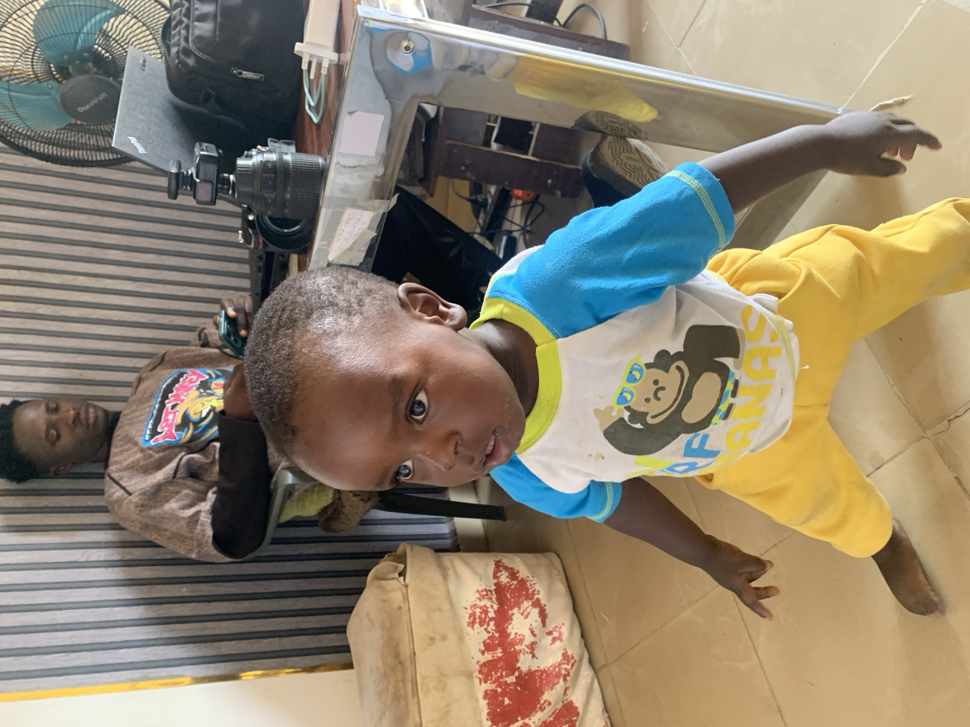 An indoor scene, likely in a home or office in Jos, Nigeria, features a young child and an adult male. The child, appearing to be a boy, is in the foreground, standing and looking directly at the camera with a neutral expression. He wears a white t-shirt with blue sleeves and a yellow collar, featuring a cartoon monkey wearing sunglasses. He also wears yellow pants.
In the mid-ground, an adult male is seen lying back or reclining in a chair or sofa with horizontal striped upholstery, engrossed in his phone. His face is visible but his expression is unclear due to the angle. He is wearing a brown long-sleeved top with a colorful graphic design on the chest.
The setting includes a light brown tiled floor and a wall with horizontal grey and white stripes. To the left, a blue-bladed "DuraVolt" fan stands near a black backpack and a closed black laptop on what appears to be a desk or table. A camera mounted on a small stand is also on this surface. The table has a reflective, possibly chrome, frame. Below the table, near the child, is a light-colored bag or sack with red markings. The overall atmosphere suggests a casual, everyday moment.