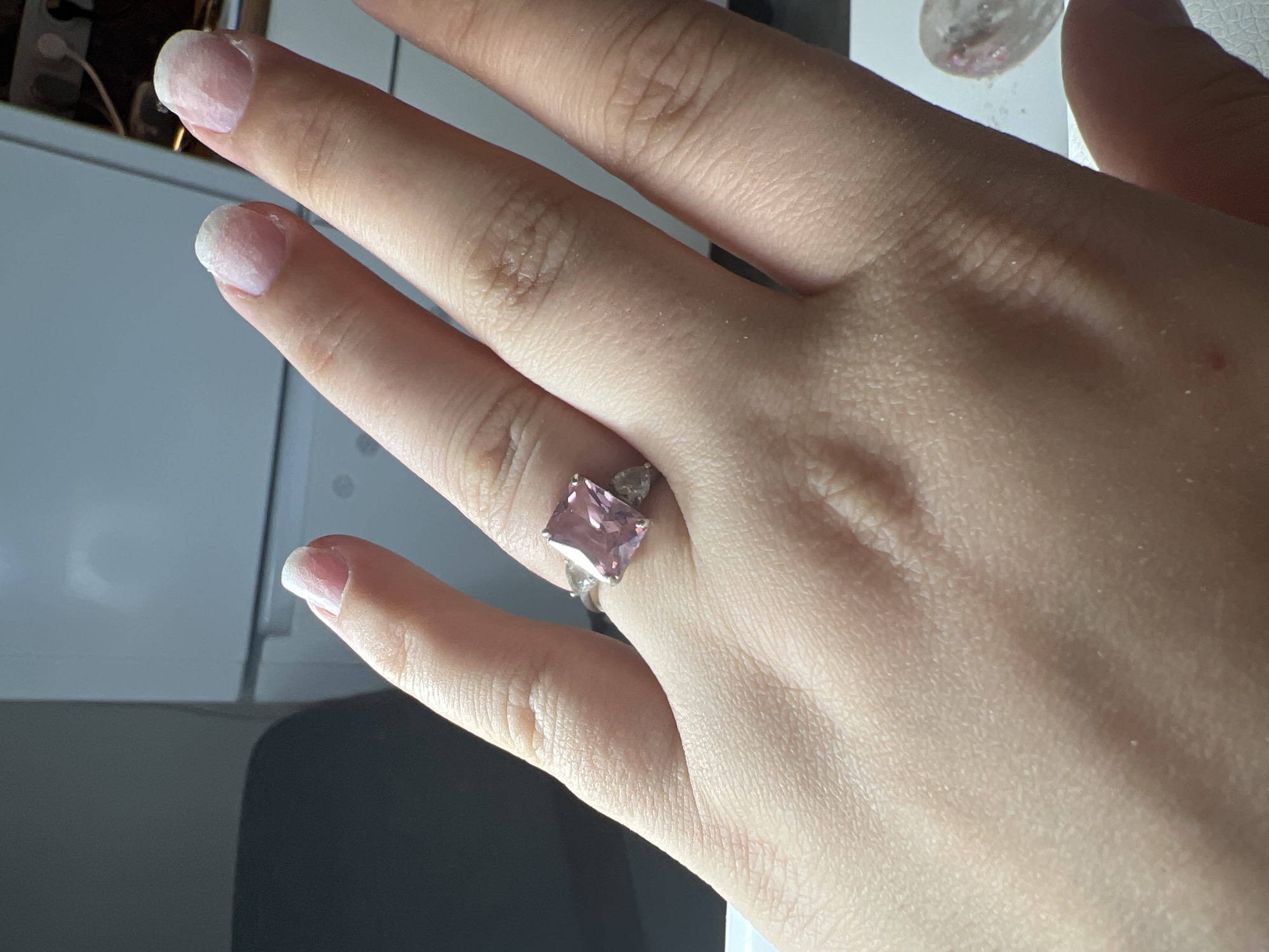 A close-up shot shows a person's left hand, adorned with a ring featuring a prominent pink rectangular gemstone. The gemstone is flanked by two smaller clear stones. The person's fingernails are visible, with a light pink gradient and white tips, suggesting a French manicure. The background is out of focus, but appears to be an indoor setting with a light-colored surface and some indistinct objects. There is no identifiable connection to Yekaterinburg, Russia, or any specific city.