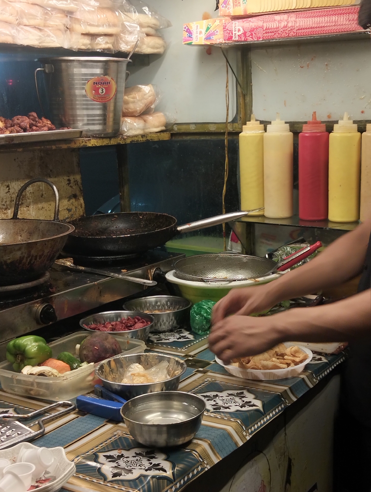 The image captures a street food preparation scene in Dhaka, Bangladesh. A vendor is seen preparing food on a counter with a patterned blue and white tablecloth. The counter is laden with various ingredients and utensils, including a clear plastic container filled with cut vegetables (green pepper, mushrooms, carrots, beetroot) and several metal bowls containing what appears to be cut meat and other prepared components. A bowl containing water sits towards the front of the counter. The vendor's hands are visible, suggesting they are actively preparing food, perhaps a street snack involving fried chips displayed in a white plastic bowl. Cooking utensils such as a grater and staple are present.
Behind the preparation area, a gas stove is visible with used cooking pans on top. The backdrop features shelves stocked with food supplies, including stacks of packaged bread. Plastic squeeze bottles filled with condiments, yellow and red, are arranged on a shelf. A stainless steel container with a logo is also noticeable on the upper shelf, indicating the presence of food storage or handling. The overall environment suggests a makeshift or semi-permanent setup typical of street food vending in Dhaka.