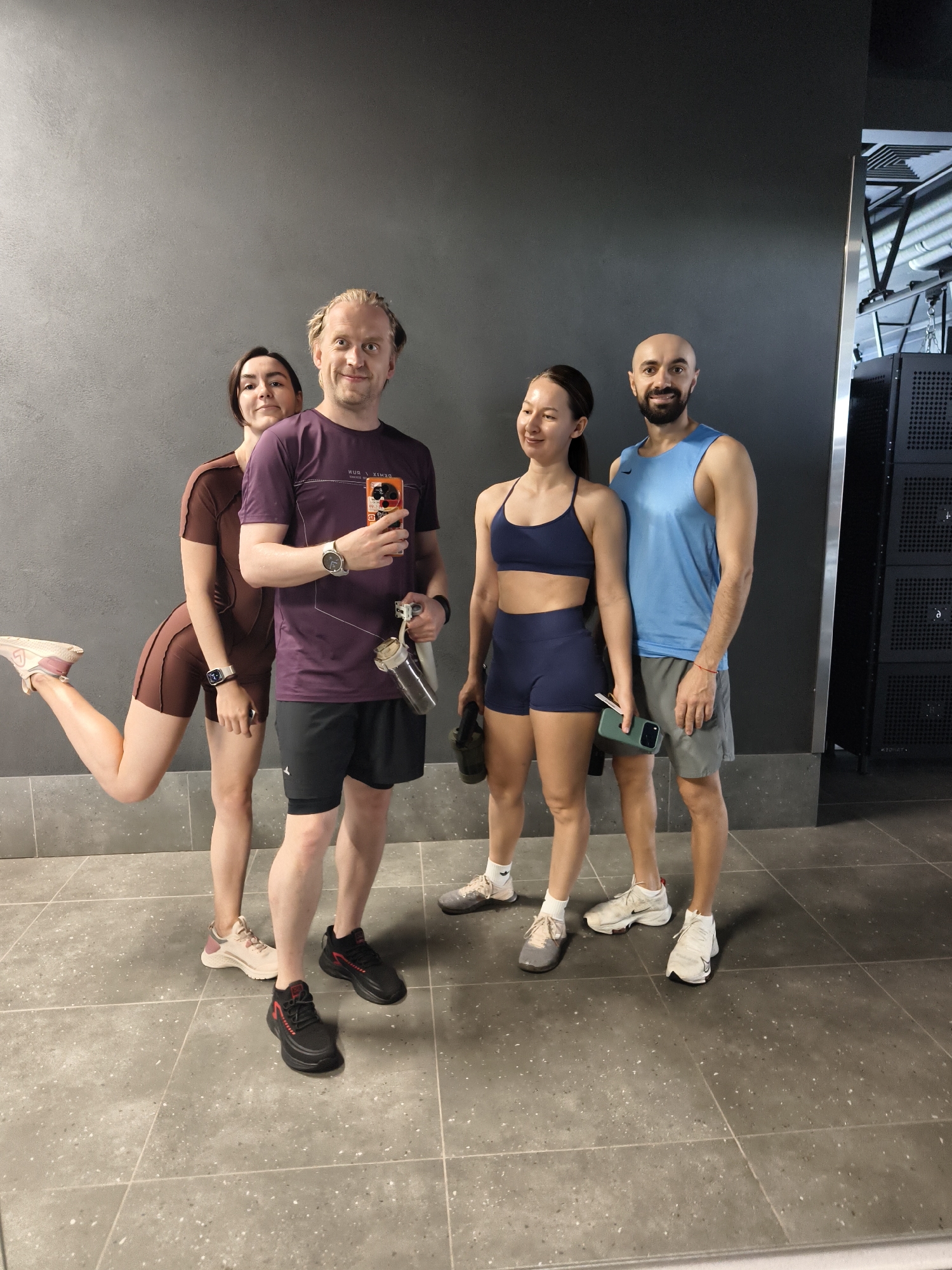 Four individuals are posed for a photograph within a modern fitness facility, likely a gym, indicated by the attire and backdrop. The setting includes a plain dark grey wall and a speckled light grey tiled floor. The "DEMIX RUN" text visible on one individual's shirt suggests a Russian context, though the specific location within Tyumen, Russia, cannot be definitively identified from the visual information.
From left to right:
A woman with short dark hair and a light complexion stands partially obscured behind the first man. She wears a form-fitting brown short-sleeved top and matching brown cycling shorts. Her right leg is lifted and bent at the knee in a playful stance, with her foot off the ground. A white smartwatch is visible on her left wrist.
Next, a man with blonde hair and a light complexion, sporting a light beard, stands centrally. He wears a purple short-sleeved athletic t-shirt, emblazoned with the inverted text "DEMIX RUN", and black athletic shorts with an integrated undershorts layer. He holds an orange-cased smartphone in his raised right hand, pointed towards the camera, and a clear water bottle with a white strap in his left hand. A silver smartwatch is on his left wrist. He is smiling directly at the camera.
To his right, a second woman with dark hair pulled back and a light complexion stands smiling gently towards her left. She wears a dark blue athletic bra top and matching dark blue athletic shorts. She holds a green-cased smartphone in her left hand and a dark, indistinct object, possibly a kettlebell, in her right hand, which is mostly out of frame.
Furthest to the right, a second man with a bald head, dark beard, and light complexion stands smiling towards the camera. He wears a light blue sleeveless athletic tank top with a small Nike swoosh logo on the left chest, and grey athletic shorts. A thin red string bracelet is visible on his left wrist.
The group appears to be in good spirits, posing collectively. The background consists of a uniformly dark grey wall. To the far right, a dark vertical structure with square vents or perforations, possibly equipment racks or lockers, is partially visible. The floor is composed of large, light grey square tiles with dark grey speckles and visible grout lines. Overhead lighting casts soft illumination on the scene.
