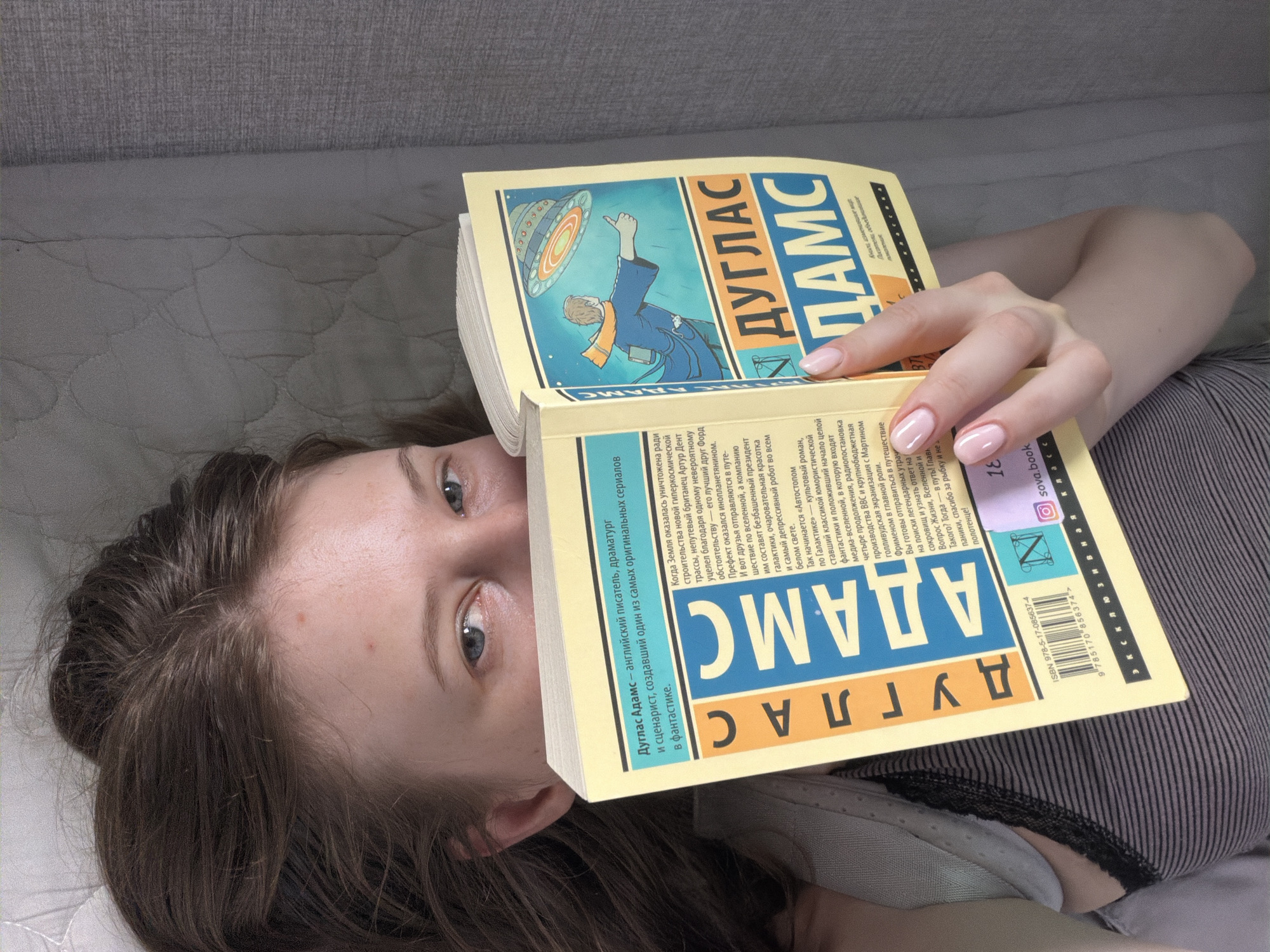 A young woman with dark hair and blue eyes is lying on her back, holding a yellow book open in front of her face. The book is titled "ДУГЛАС АДАМС" in large blue letters and features an illustration of a flying saucer. She is wearing a grey and black striped tank top. The background appears to be a textured grey surface, possibly a bed or sofa. The image is taken from a high-angle, slightly overhead perspective. No specific location within Dongducheon-si, South Korea is identifiable.