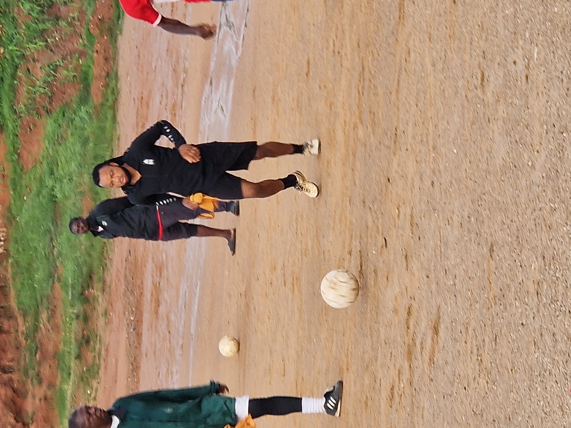 The image shows a group of men on a dusty, outdoor sports field. The ground is a mixture of dry earth and patches of wetness, possibly from recent rain, with some sparse green vegetation in the background. In the foreground, a man in a black athletic shirt and shorts is dribbling a soccer ball. He is holding a yellow cloth in his left hand. To his left, another man in a dark green jacket and black leggings is standing, with a soccer ball near his feet. Further back, two other men in athletic wear are visible. One is wearing a black jacket over a red shirt, and the other is partially visible in a red shirt. The scene appears to be a casual soccer game or practice session in Kabong, Nigeria.