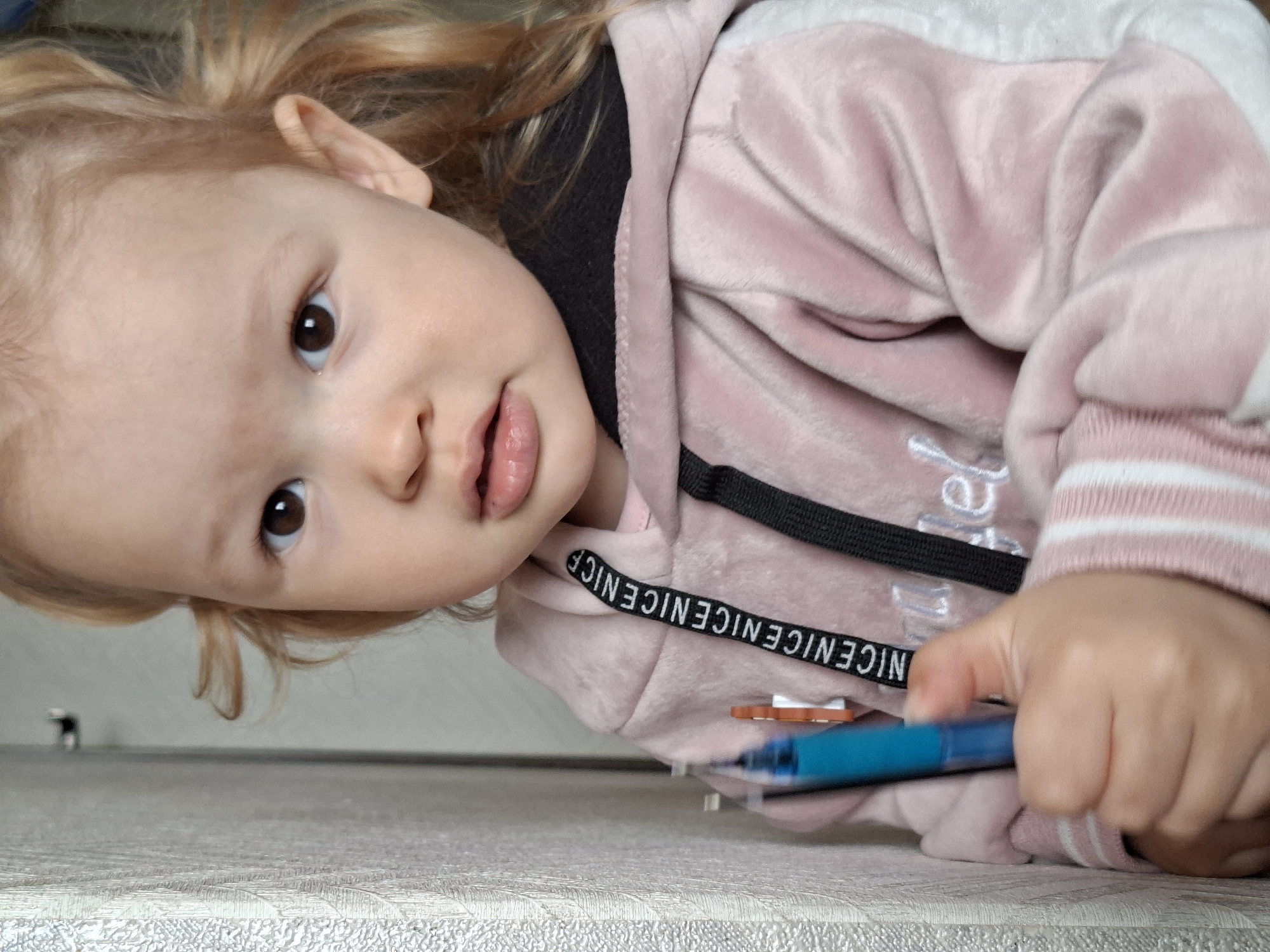 A child with fair skin and light blonde hair is lying on a textured gray surface, possibly a table or floor. The child is wearing a pink hooded sweatshirt with white and pink striped trim on the sleeves. The hood is lined with a black fabric, and the drawstrings of the hood have the word "NICE" printed repeatedly along their length. The child is looking directly at the camera with brown eyes and a slightly open mouth. In their right hand, they are holding a blue pen or marker. The location is unknown.