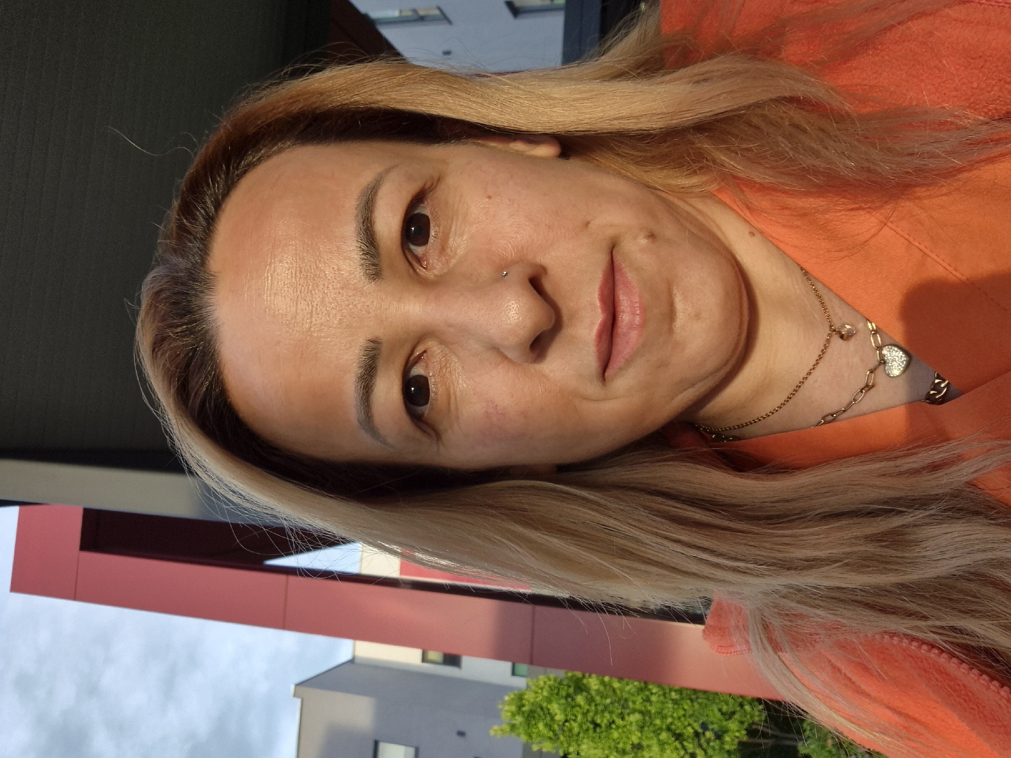 A medium shot captures a woman in her late 30s or early 40s, facing the camera directly. She has medium-length blonde hair, parted to her left, and fair skin. She is wearing an orange hooded jacket and a gold chain necklace with a heart-shaped pendant. A nose ring is visible on her right nostril.
The background is outdoors, with modern buildings and a bright red architectural feature to her left. A small, green bush is also visible to her left. The lighting suggests it is late afternoon or early evening, with the sun casting a warm glow. The environment appears to be an urban or suburban setting.