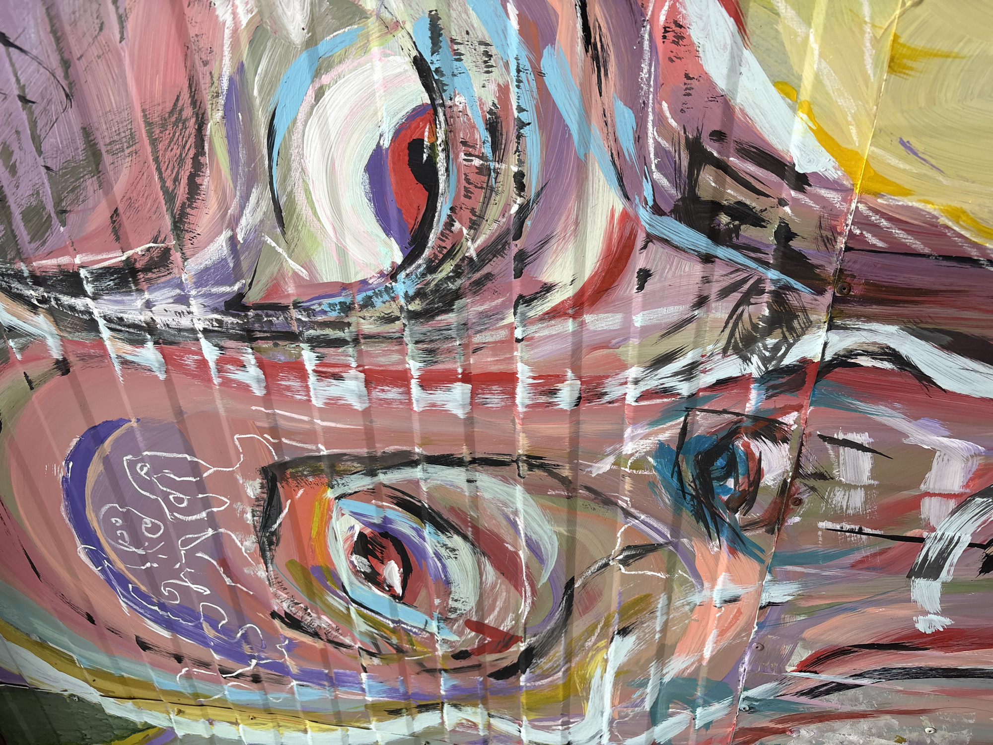 The image shows a close-up of painted corrugated metal. The painting features abstract depictions of two fish or aquatic creatures, identifiable by circular areas suggesting eyes. The color palette consists of pinks, purples, blues, white, yellow, and black. The style of painting is expressive, with visible brushstrokes. The image appears to be a section of a larger mural, possibly public art in Kragujevac, Serbia, judging by the image's origin. The corrugated metal serves as the canvas, adding a textured background to the artwork.