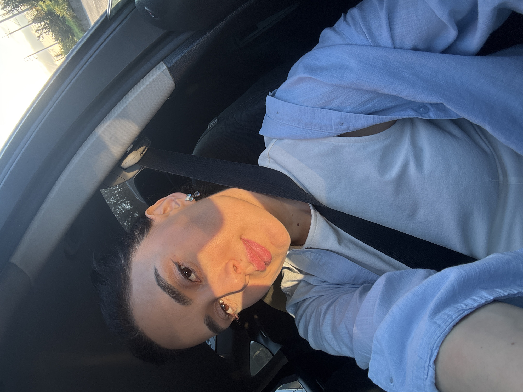 The image, rotated vertically, captures a head-and-shoulders selfie of a young adult individual inside a car, likely in the driver's or front passenger's seat. The person has dark hair, pronounced dark eyebrows, and dark eyes, looking directly at the camera with a slight smirk. Their lips are painted with a pinkish-red lipstick, and they wear an earring with small, clear stones in their visible left ear.
They are dressed in a light blue long-sleeved button-up shirt worn over a white t-shirt, with a black seatbelt fastened across their chest. Intense, warm sunlight, characteristic of early morning or late afternoon, illuminates the right side of their face and the car's interior, creating a contrast with the shadowed areas.
Through the car's side window, a sunny outdoor environment is visible, featuring trees, a road, and what appear to be utility poles. The car's interior details, including parts of the dashboard, A-pillar, and a headrest, are discernible. The word "AIRBAG" is faintly visible on a panel in the car's interior. The setting is Tbilisi, Georgia.