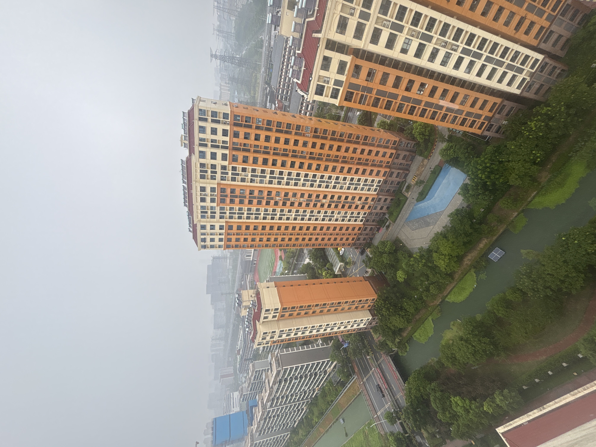 This is an aerial, high-angle view of a modern urban residential area in Suzhou Industrial Park, China, under an overcast sky. The scene is dominated by several tall apartment buildings with distinct orange and cream facades, featuring numerous windows. In the foreground, a green, algae-covered canal runs through the landscape, bordered by lush trees. A red bridge spans the canal in the midground. To the left, a row of longer, lower apartment buildings with white and grey facades stretches into the distance. In the background, the cityscape recedes into a hazy horizon, punctuated by industrial structures, power lines, and more buildings. A blue, irregularly shaped swimming pool is visible in the courtyard of one of the apartment complexes. No people are discernible in the image.