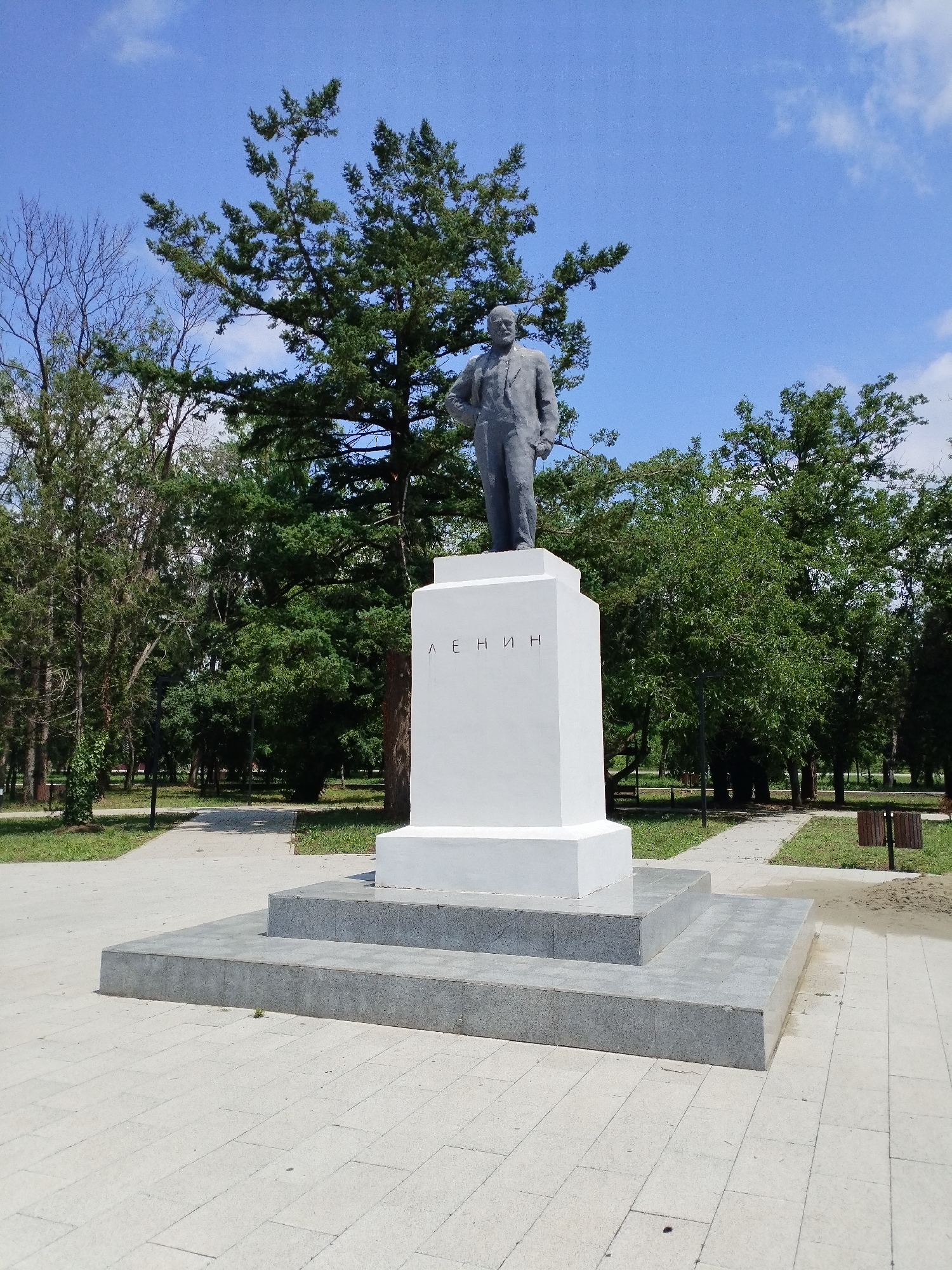 The image shows a statue of Vladimir Lenin in Vinogradny, Russia. The statue is made of gray stone and stands on a white pedestal. The pedestal has the name "LENIN" inscribed on it in Cyrillic letters. The statue is located in a park with trees and a blue sky. The area around the statue is paved with gray tiles. The overall scene depicts a monument in a public park.