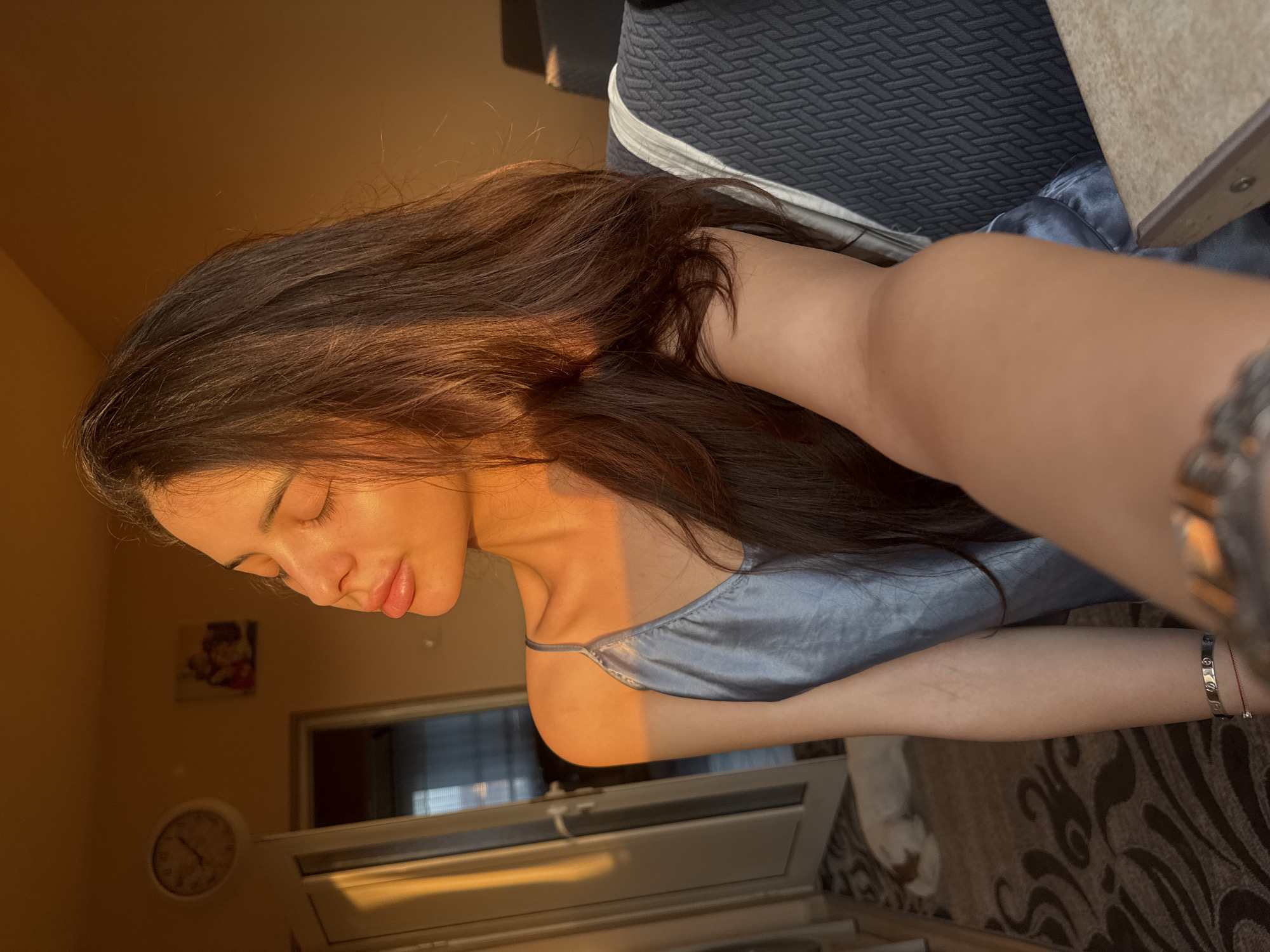 The image displays an indoor setting, illuminated by warm, directional sunlight entering from the left. A female individual, seen from the chest up, occupies the central and right portions of the frame. Her eyes are closed, and her head is tilted upwards, with strong golden light illuminating the right side of her face and her long, dark brown hair. She is wearing a light blue or greyish-blue silky camisole. On her left wrist, she wears a silver-toned bracelet and a red string bracelet. Her right arm is extended towards the camera, indicating a selfie.
In the background, on the left, a light-colored wall features a white circular analog clock and a framed picture. Below these, a white door frame leads to another room with a partially visible window and blinds. On the floor, a brown and beige patterned rug is visible, upon which a small, light-colored dog is lying down, out of focus. To the right of the female, a dark grey or black upholstered chair or sofa with a textured pattern and a white stripe detail is present. Part of a light-colored tabletop or surface is visible in the extreme foreground on the right. The image does not contain visual information to forensically confirm the location as Plovdiv, Bulgaria.